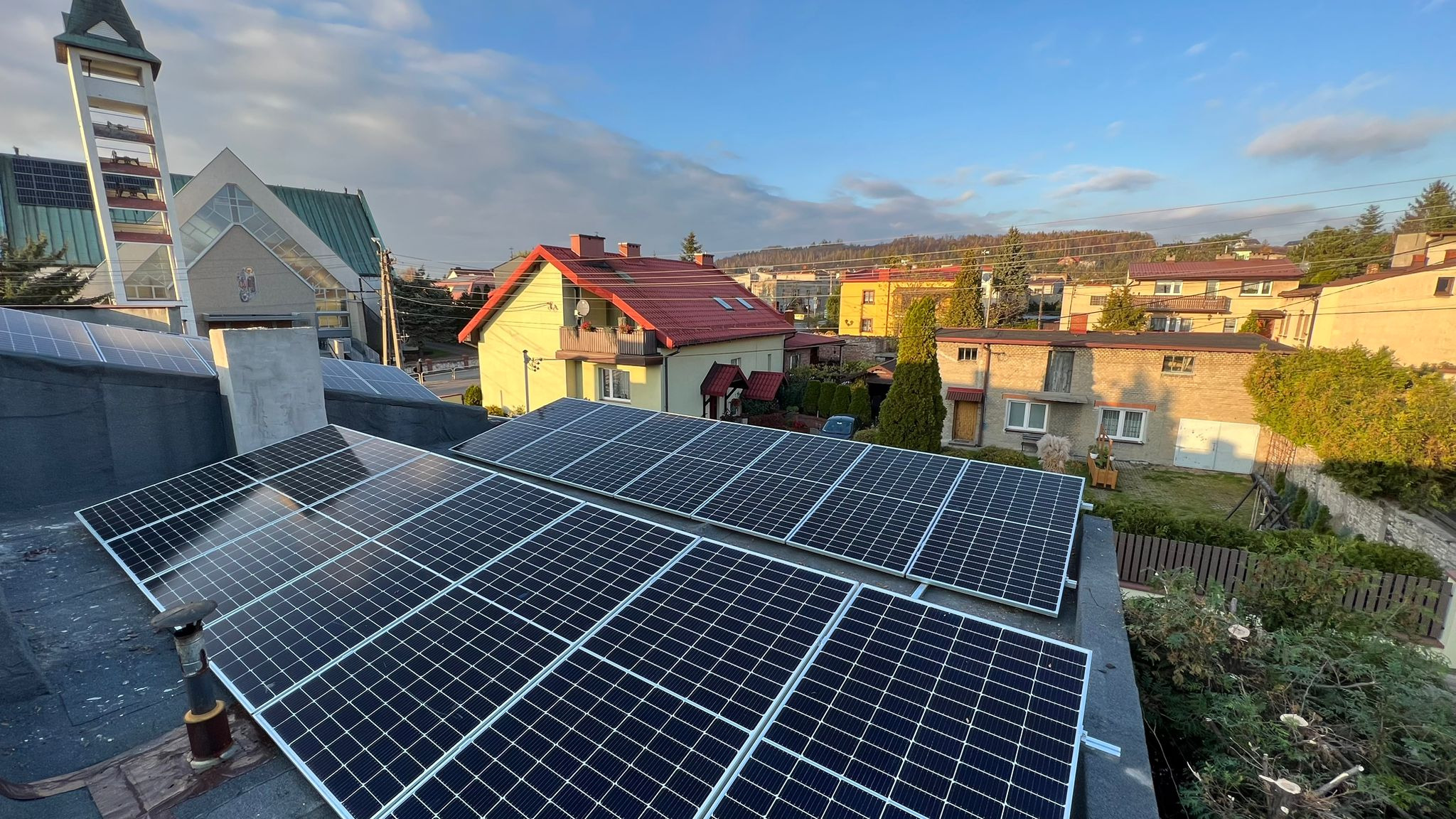 Panele słoneczne na dachu budynku mieszkalnego, widok z góry na miasto i kościół w tle, ciepłe światło popołudniowe.