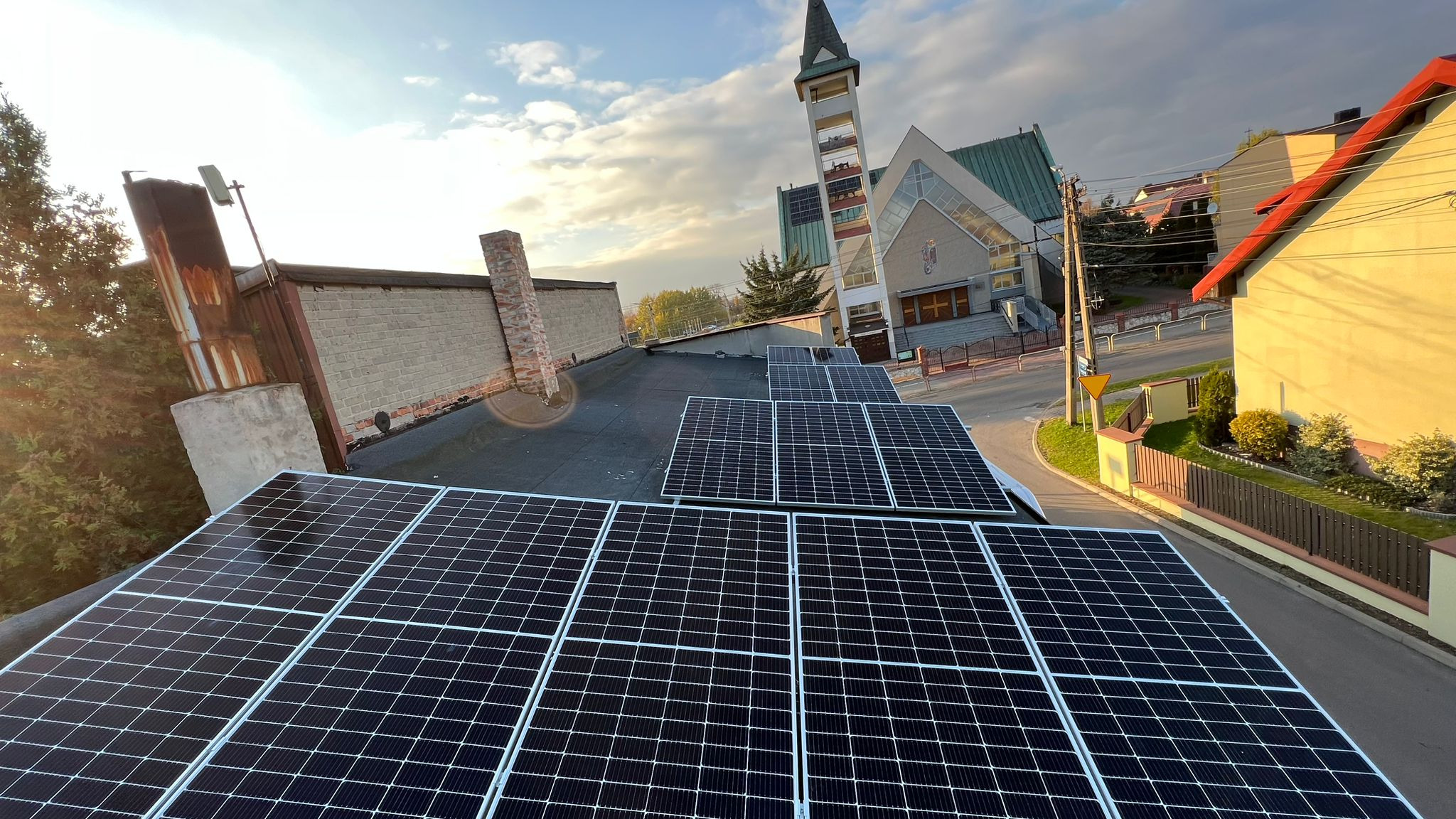 Instalacja paneli słonecznych na płaskim dachu budynku z widokiem na kościół z wieżą w tle, w ciepłym świetle popołudniowego słońca.