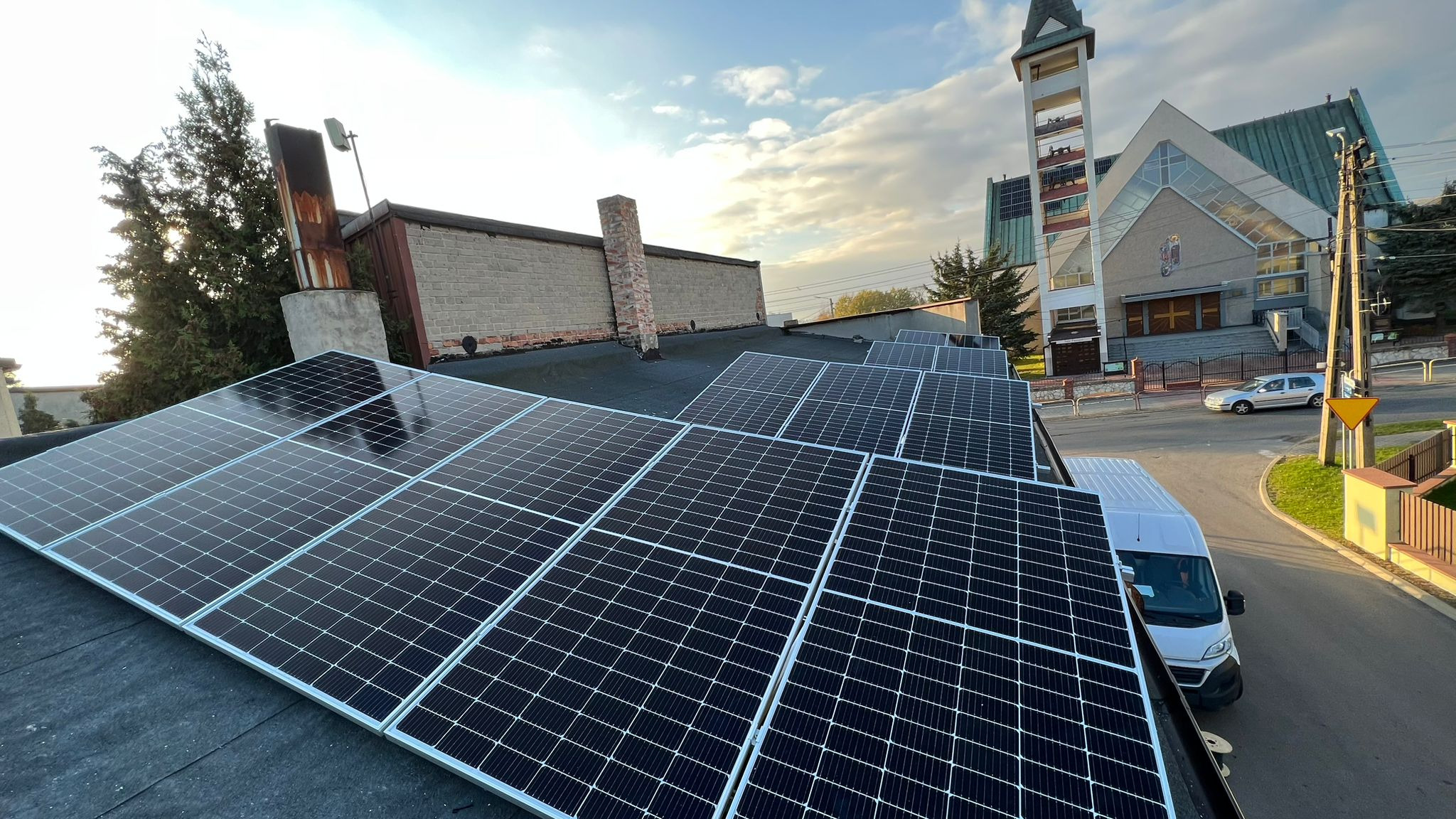 Panele słoneczne na dachu budynku z kościołem w tle, widok z góry, biały samochód dostawczy zaparkowany obok.