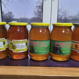 Pięć słoików z miodem różnego rodzaju (lipowy, z terenów leśnych, słonecznikowy) od Pasieki Pszczela Białowieska Barć, ustawionych rzędem na blacie, z widocznym oknem w tle.