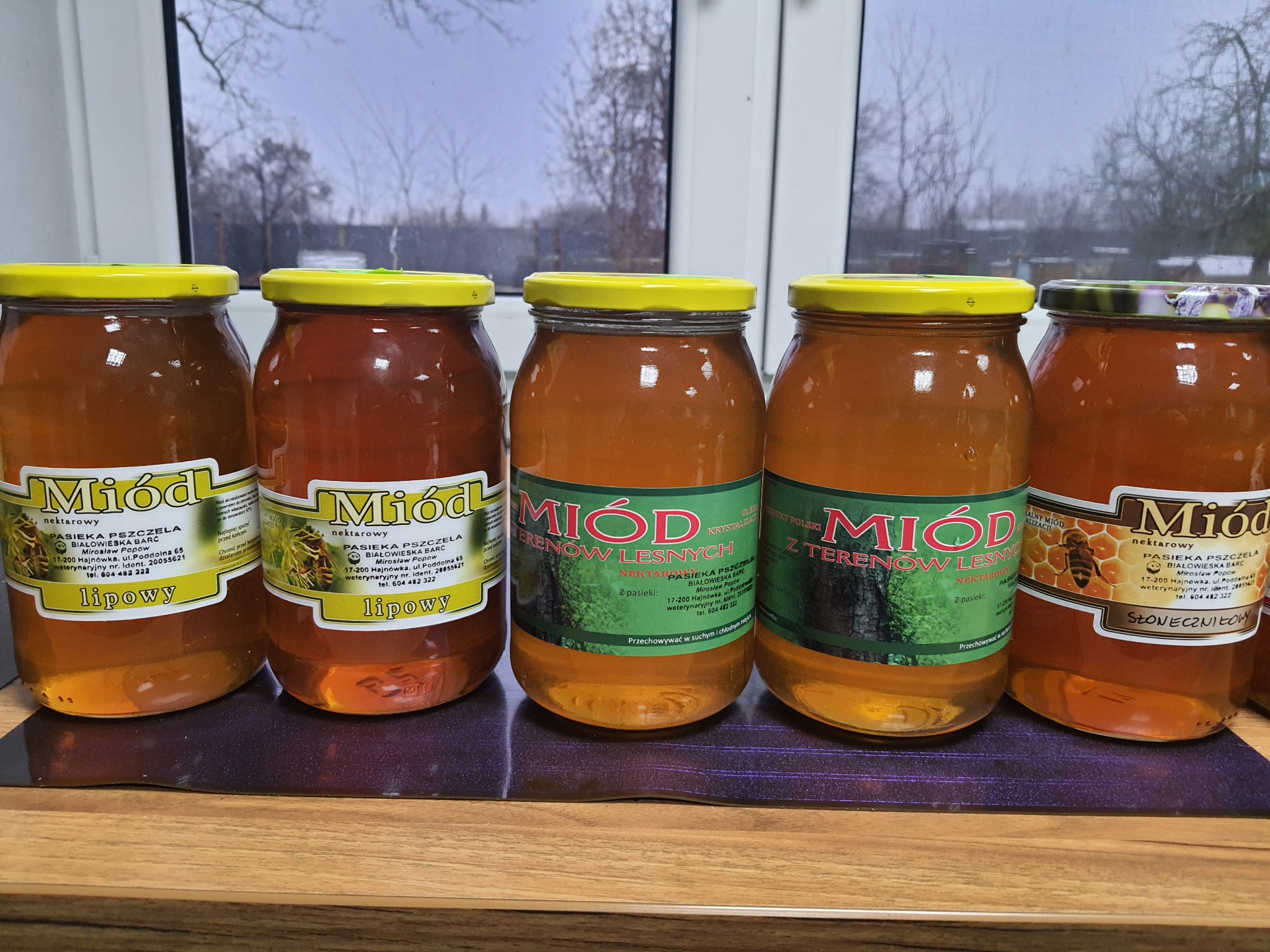 Pięć słoików z miodem różnego rodzaju (lipowy, z terenów leśnych, słonecznikowy) od Pasieki Pszczela Białowieska Barć, ustawionych rzędem na blacie, z widocznym oknem w tle.