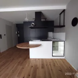 Nowoczesna kuchnia w odcieniach bieli i czerni z drewnianym blatem, widok na fragment podłogi z paneli drewnianych i minimalistyczną dekorację ścienną.