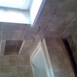 Ukończona łazienka z brązowymi, kwadratowymi płytkami ceramicznymi na ścianach i podłodze, widoczne zawory i fragment kabiny prysznicowej z białymi ramami.