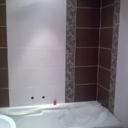 Świeżo wykafelkowana ściana nad wanną z białymi i brązowymi płytkami, dekoracyjna bordiura z motywem roślinnym, wanna zabezpieczona folią ochronną.