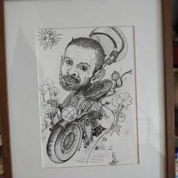 OdKarykatury DoMuralu - twoja wizja, moja pasja - Oprawiony w ramkę rysunek tuszem przedstawiający karykaturę brodatego mężczyzny na motocyklu, z elementami humorystycznymi jak słońce z zębami i drzewa z oczami, sygnowany w prawym dolnym rogu.