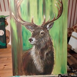 OdKarykatury DoMuralu - twoja wizja, moja pasja - Obraz na płótnie przedstawia portret jelenia na tle zielonego lasu, umieszczony na drewnianej sztaludze w pracowni artystycznej. Widoczne pędzle i farby.