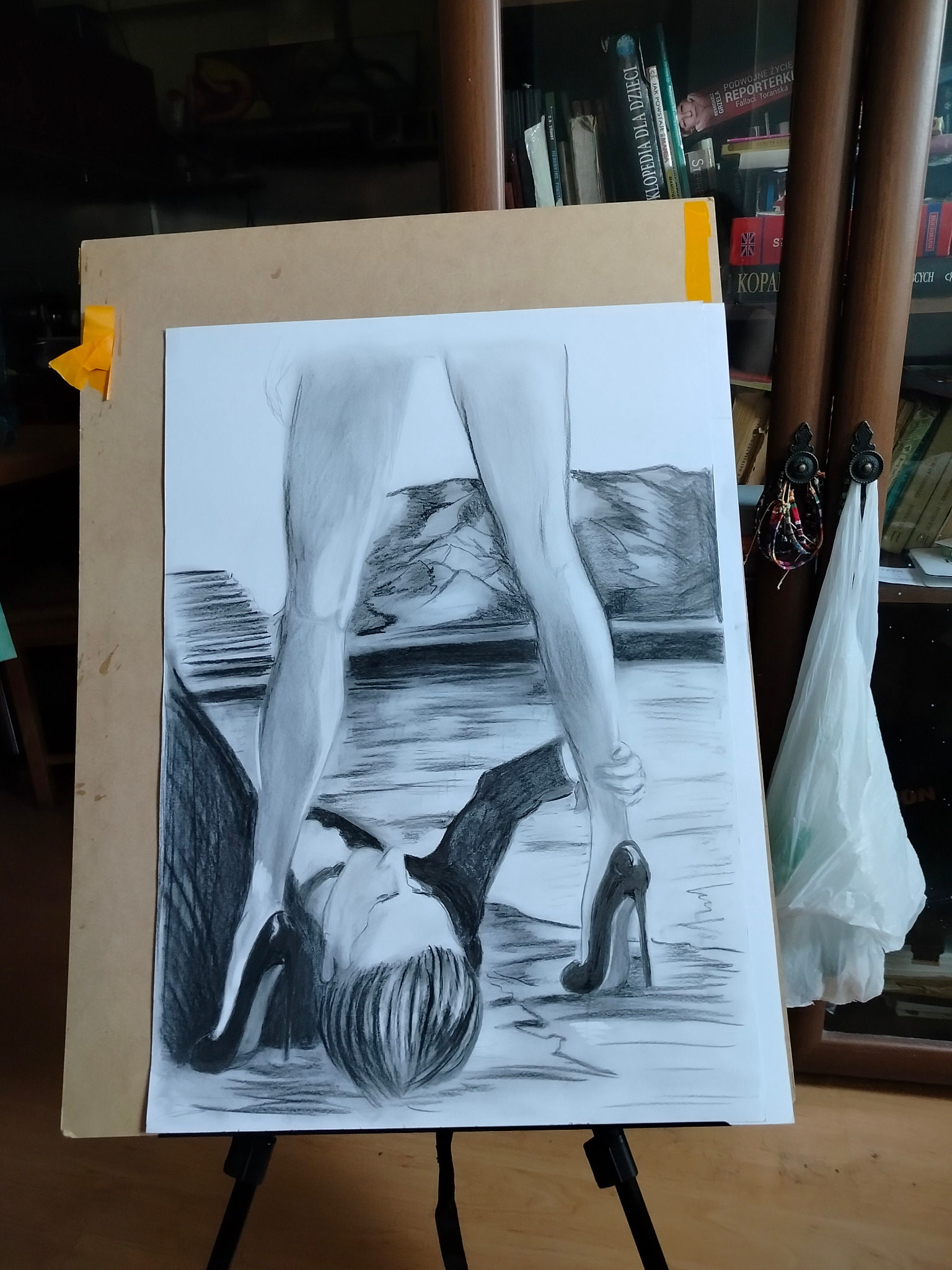 Szkic ołówkiem na papierze przedstawiający kobietę w szpilkach stojącą na leżącym mężczyźnie, umieszczony na sztaludze w pracowni artystycznej.