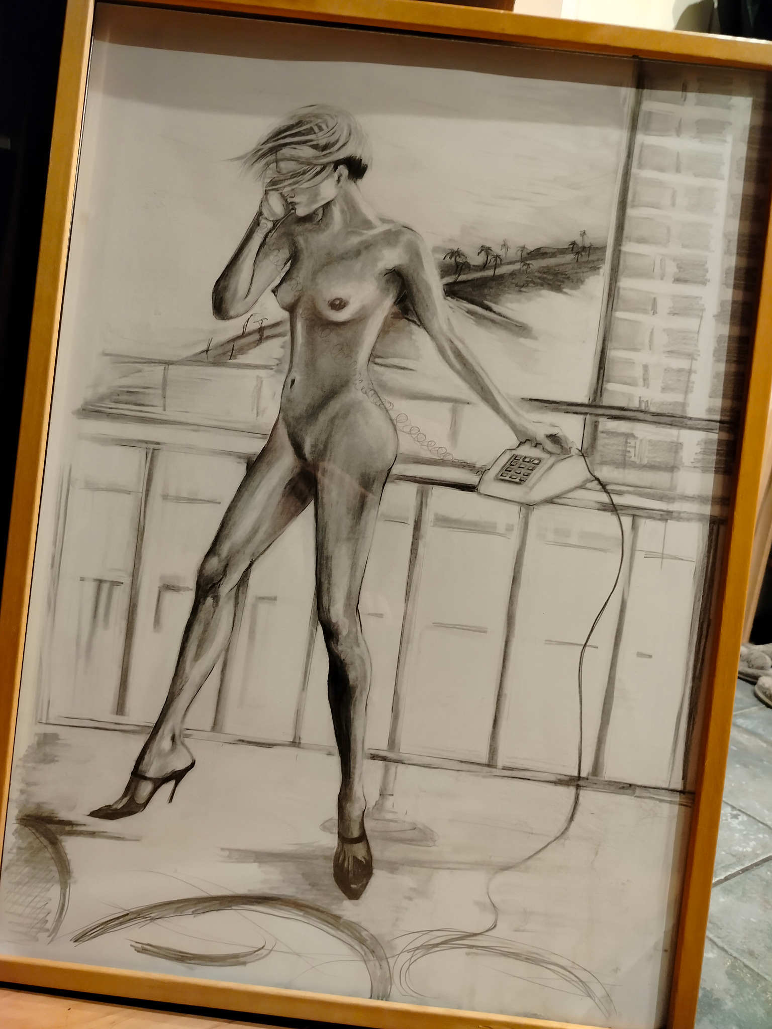 Szkic nagiej kobiety w szpilkach rozmawiającej przez telefon na balkonie z widokiem na palmy i budynek, oprawiony w drewnianą ramę.