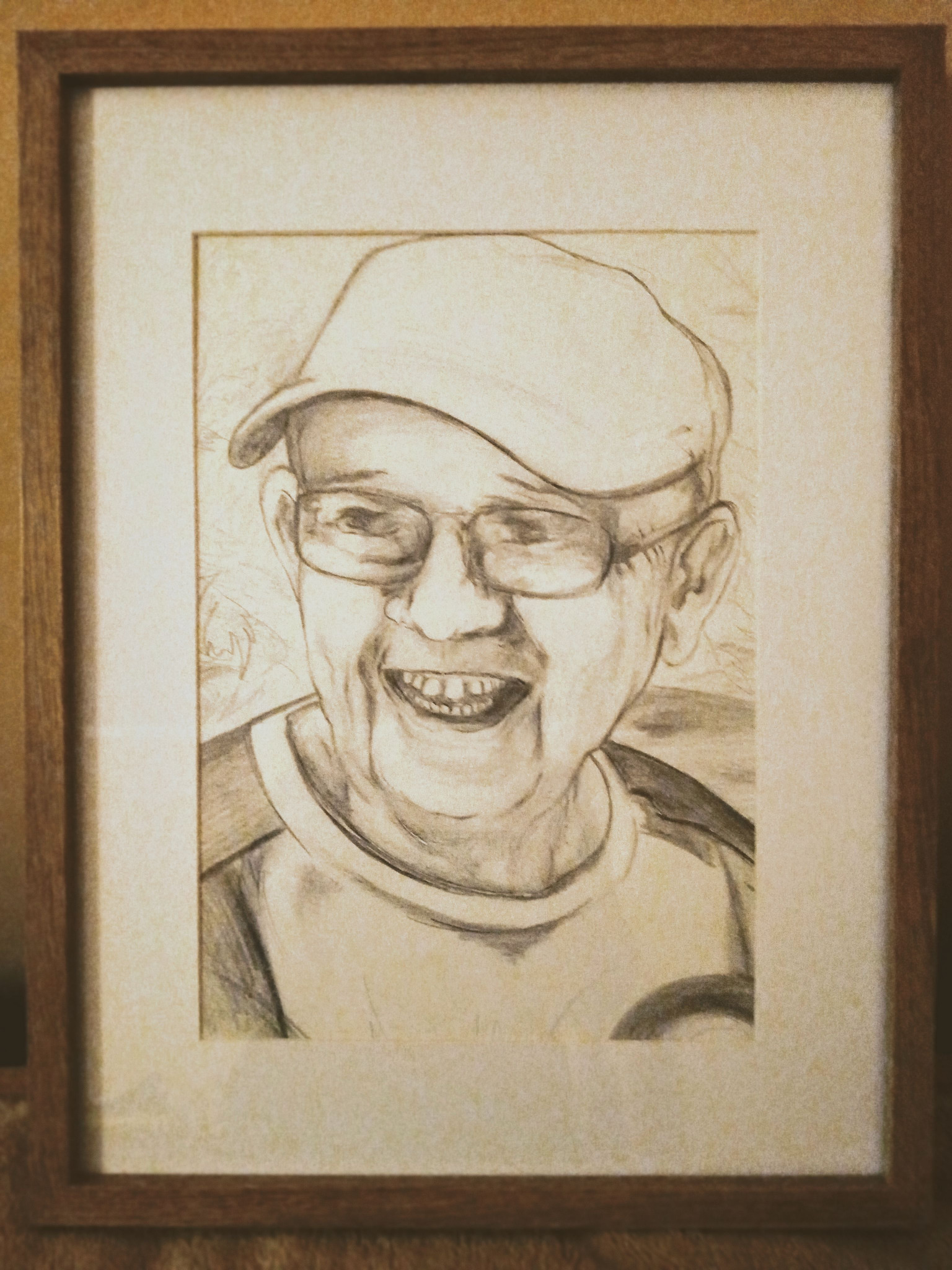 Oprawiony w drewnianą ramę portret uśmiechniętego mężczyzny w czapce i okularach, wykonany ołówkiem na beżowym papierze.