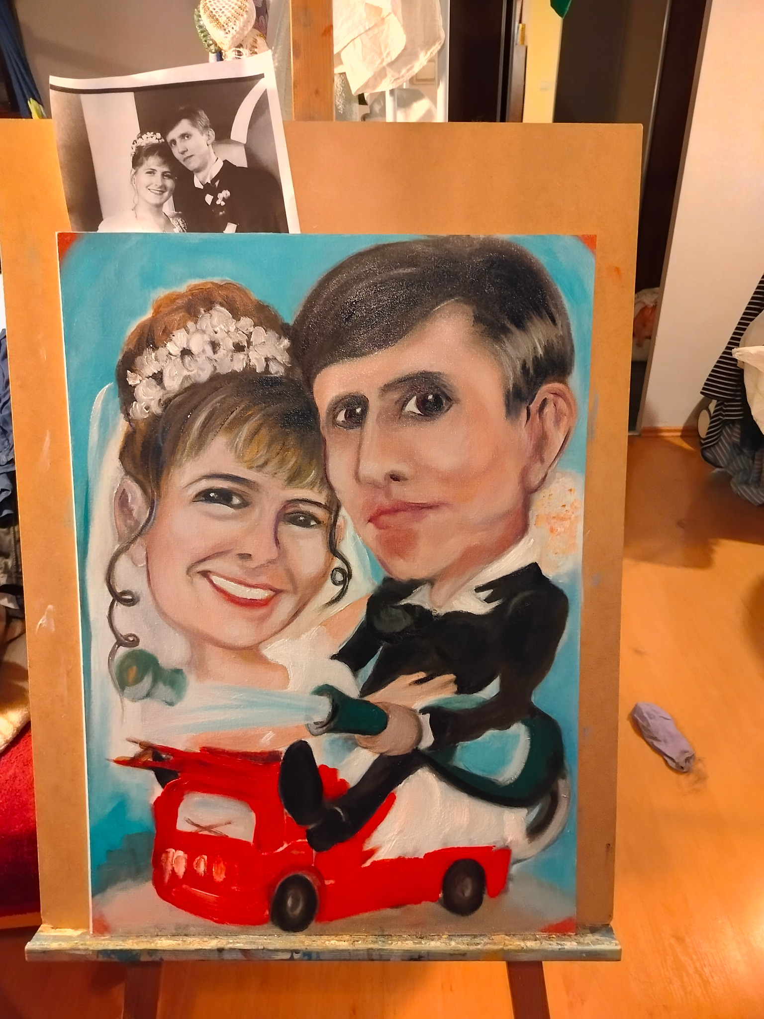 Niedokończony portret ślubny w stylu karykatury, namalowany farbami olejnymi na płótnie, przedstawiający parę młodą siedzącą w czerwonym samochodzie strażackim, obok leży fioletowa szmatka malarska.