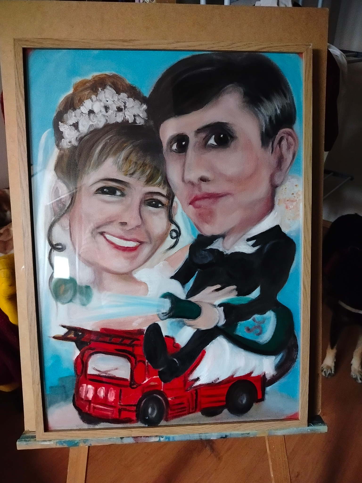 Oprawiony portret pary młodej w stylu karykatury, jadącej na czerwonym wozie strażackim na błękitnym tle.