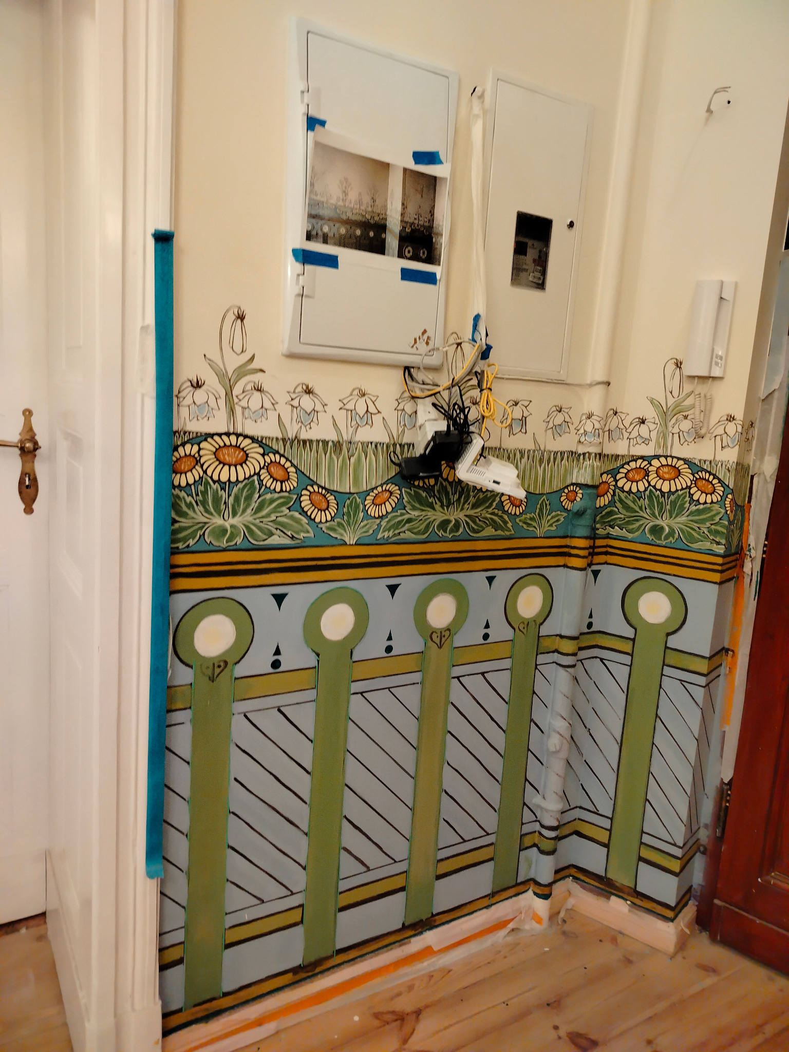 Ręcznie malowana dekoracja ścienna z motywami kwiatowymi i geometrycznymi w stylu secesyjnym, częściowo zasłonięte gniazdka elektryczne i zabezpieczenia na ścianie.