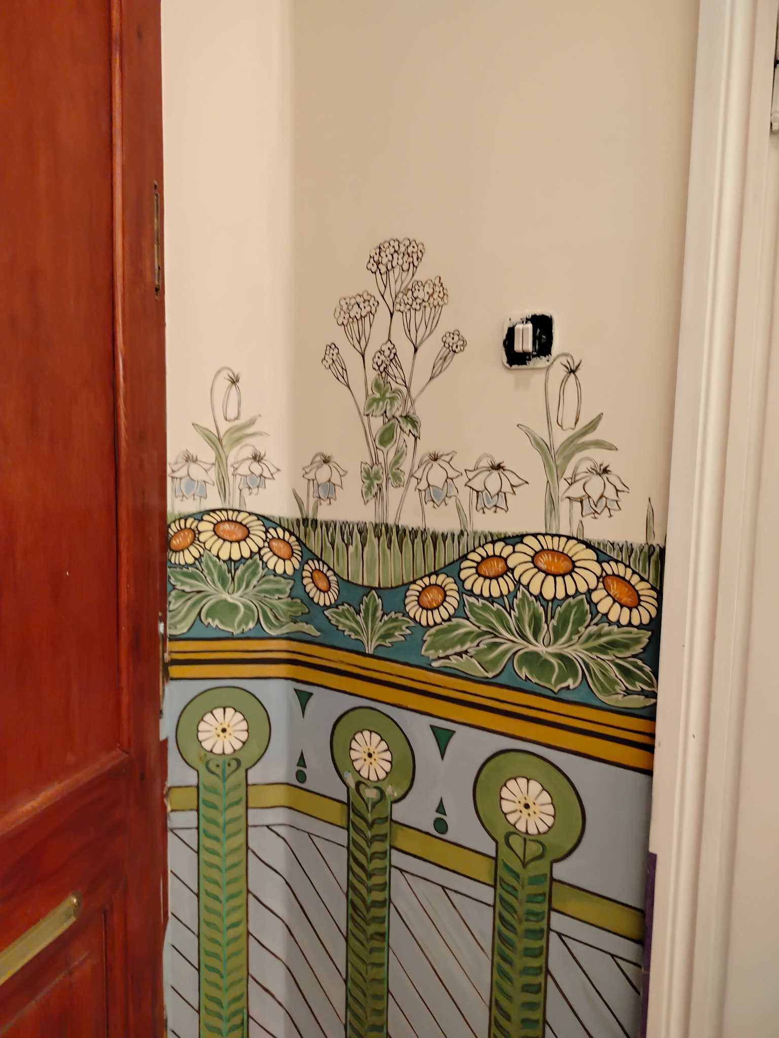 Kwiatowy motyw dekoracyjny na ścianie z widocznym włącznikiem światła, utrzymany w stylu secesyjnym z dominującymi odcieniami zieleni, błękitu i żółci, częściowo zasłonięty przez otwarte, drewniane...