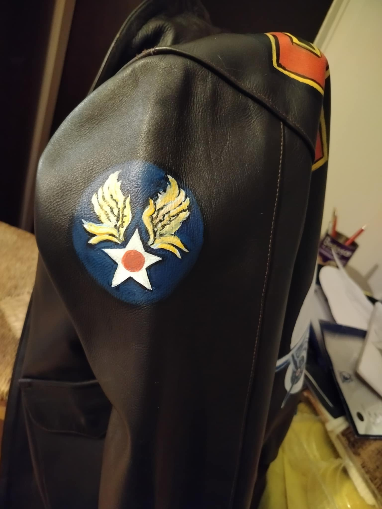 Brunatna skórzana kurtka z ręcznie malowanym motywem lotniczym: biała gwiazda z czerwonym okręgiem otoczona złotymi skrzydłami na niebieskim tle, widoczne detale szwów i kieszeń.