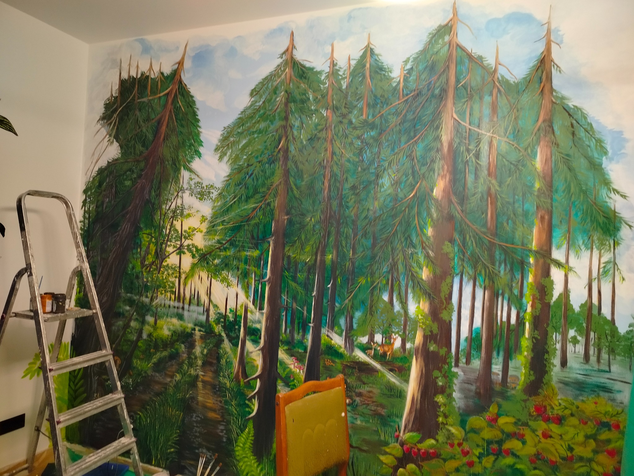 Mural na ścianie przedstawia leśny krajobraz z jeleniami, malowany farbami. Widoczna drabina malarska i krzesło, sugerujące etap pracy nad dziełem.