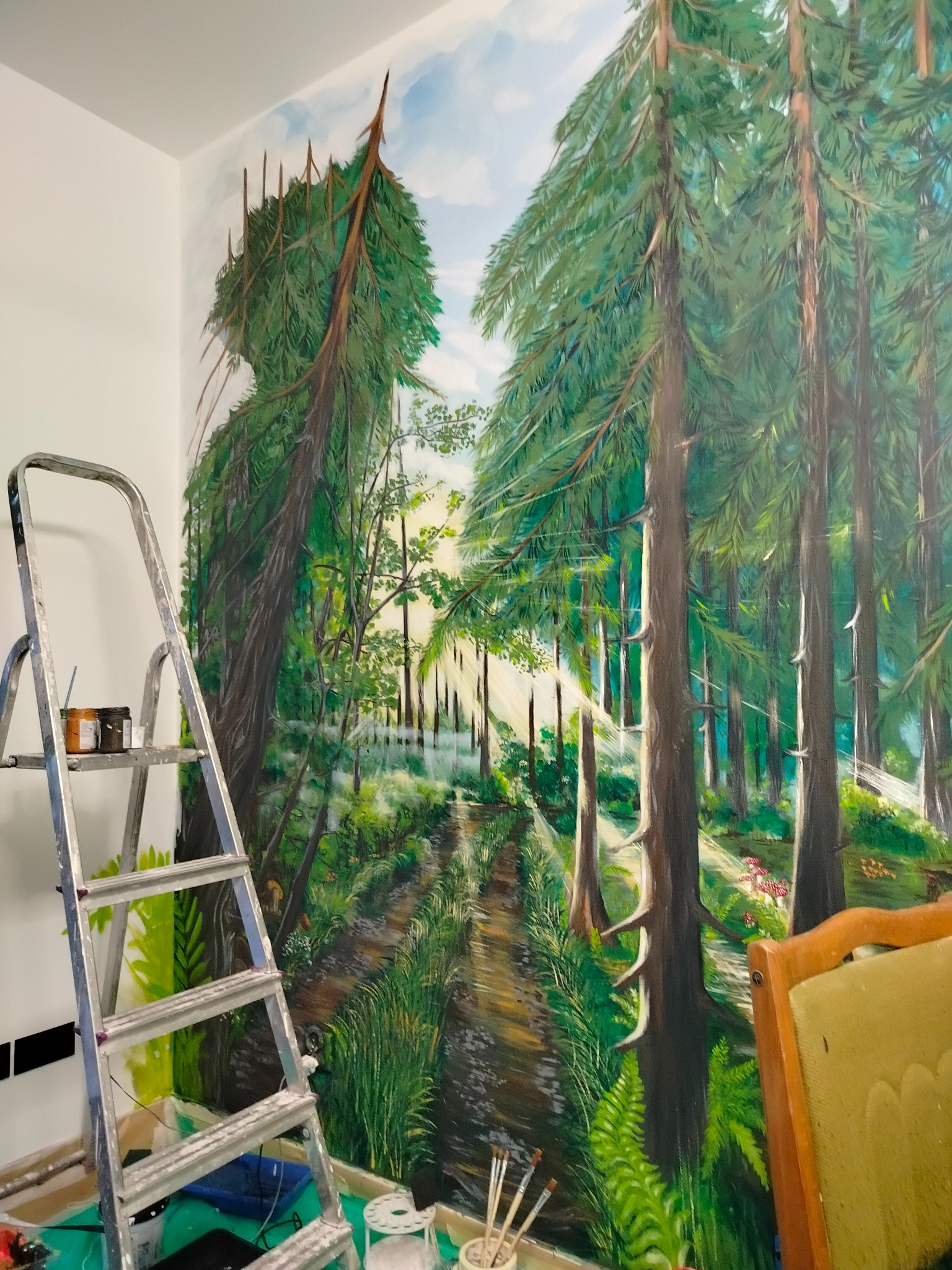 Mural na ścianie przedstawiający leśny krajobraz z perspektywą ścieżki, drabina malarska z farbami i pędzlami w pobliżu, krzesło w prawym dolnym rogu.