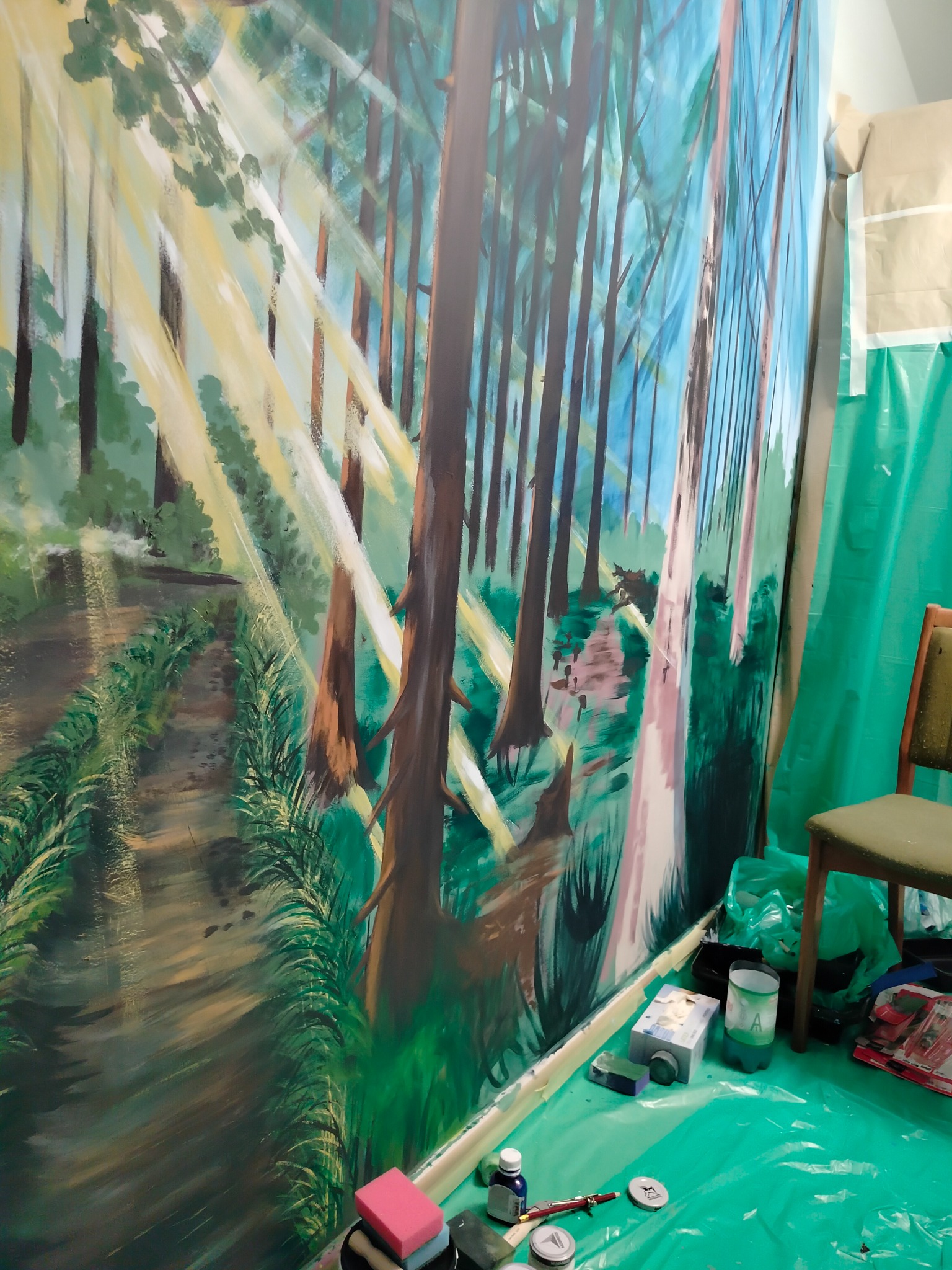 Mural leśny w trakcie tworzenia: widoczne farby, pędzle i krzesło. Promienie słońca przebijają przez drzewa, tworząc malowniczy krajobraz w zieleni i błękicie.