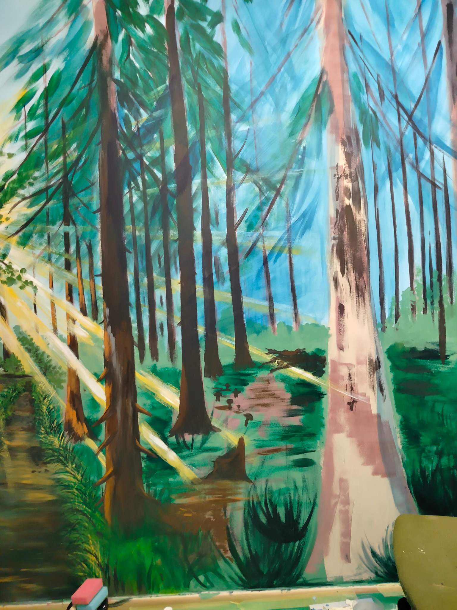 Ręcznie malowany pejzaż leśny z promieniami słońca, wykonany farbami na ścianie. Widoczne drzewa, zieleń i niebieskie niebo w tle. Artystyczna wizja lasu.