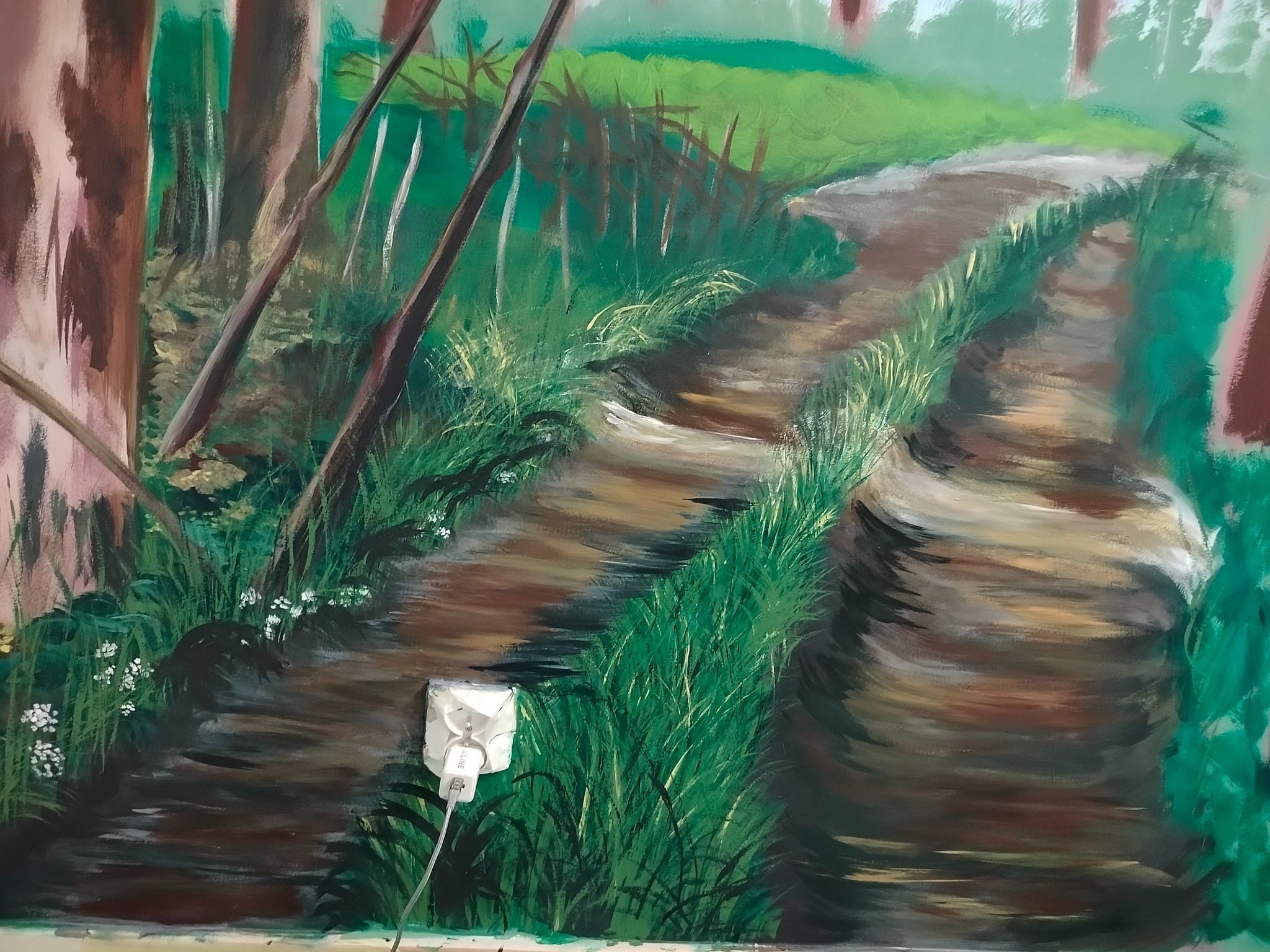 Malowidło ścienne przedstawiające leśną ścieżkę z bujną zielenią i drzewami, z widocznym gniazdkiem elektrycznym w dolnej części obrazu.