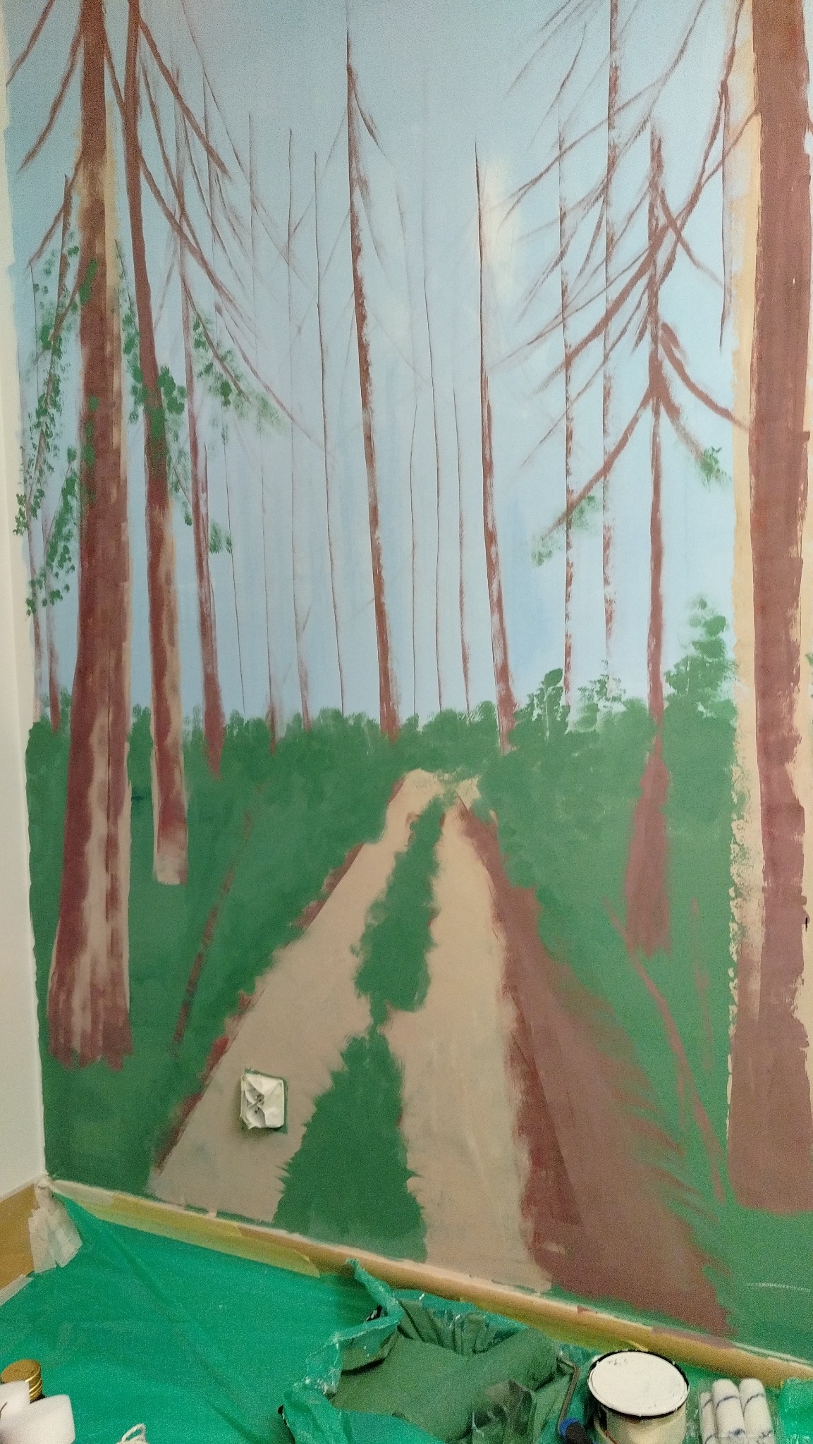 Mural na ścianie przedstawia leśną ścieżkę z wysokimi drzewami. Widoczne farby i zabezpieczenia podłogi sugerują etap tworzenia dzieła.