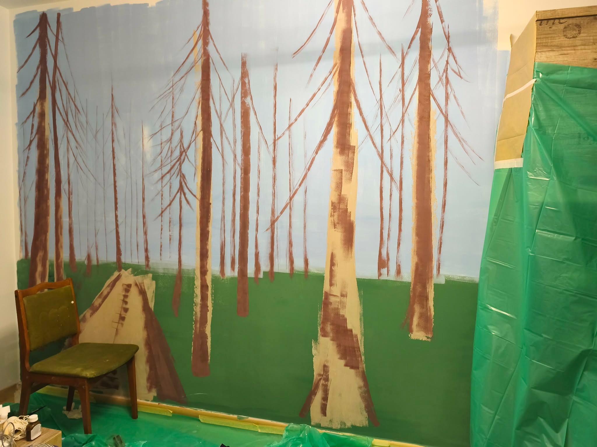Mural z motywem lasu w trakcie malowania na ścianie. Widoczne drzewa w odcieniach brązu i zieleni, niebieskie niebo. Krzesło i folia malarska sugerują etap pracy.