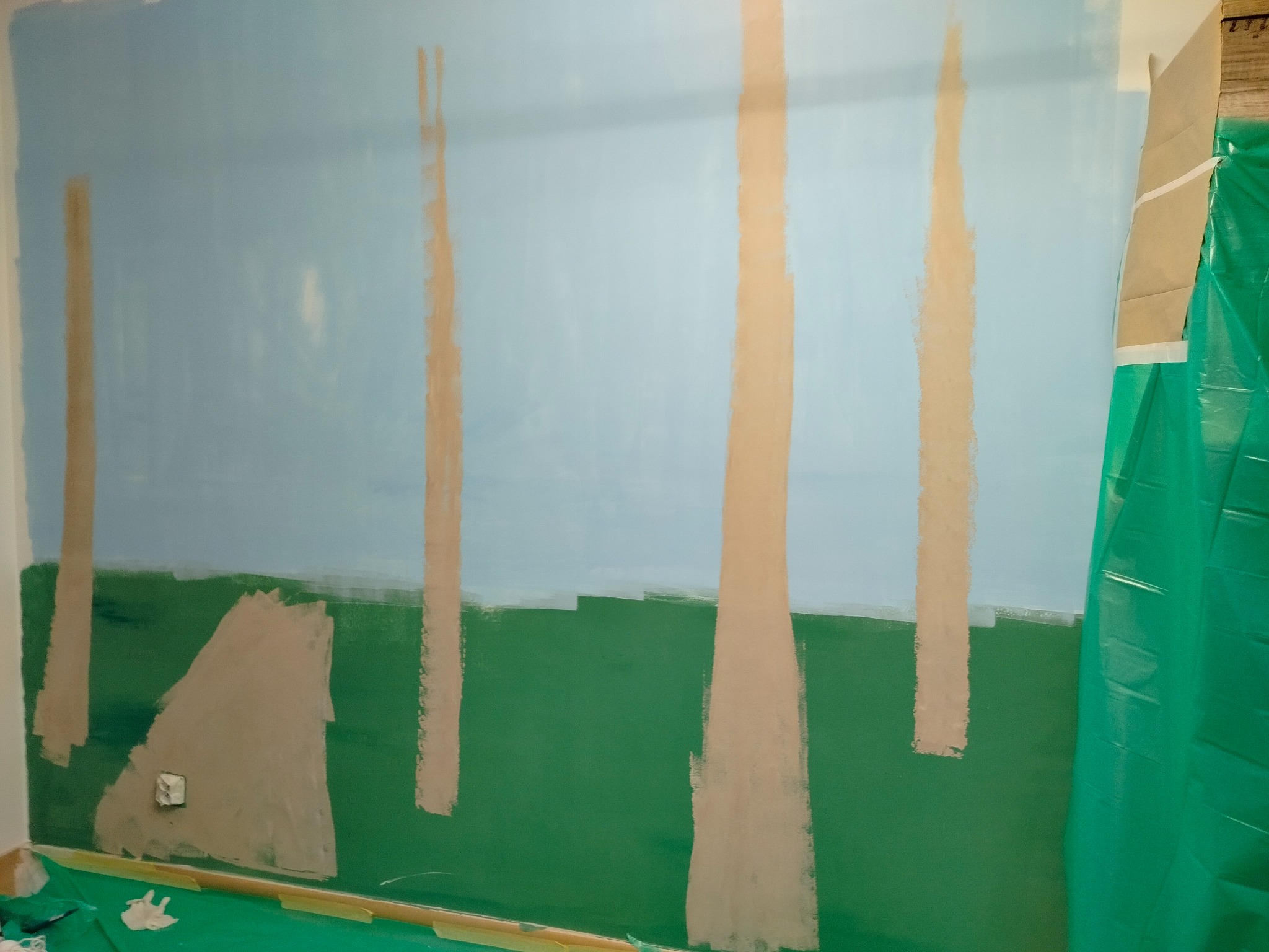 Ściana w trakcie malowania artystycznego: niebieskie niebo, zielona trawa i brązowe pnie drzew, zabezpieczona folią malarską. Widoczny etap tworzenia muralu.