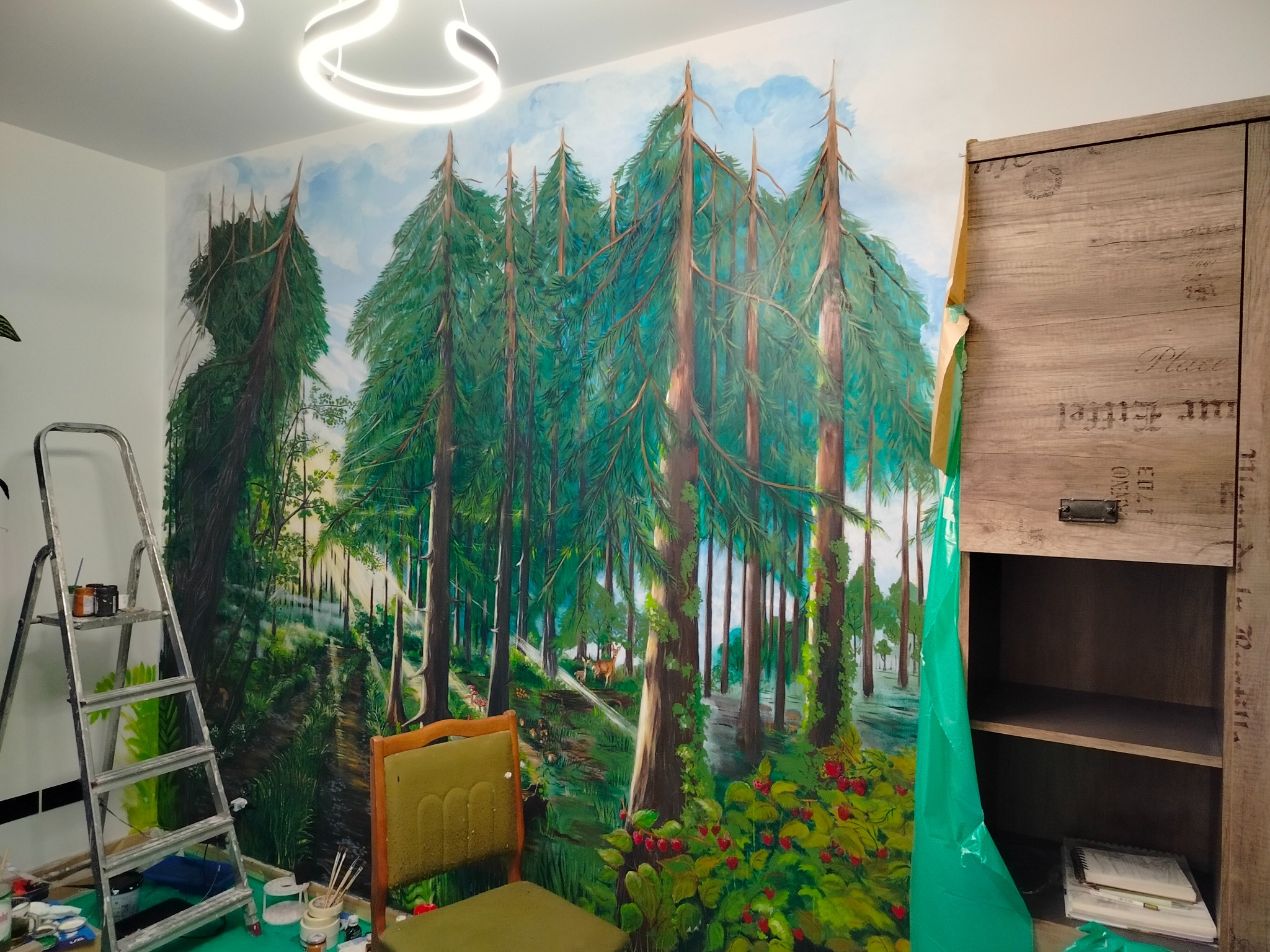 Mural z motywem leśnym na ścianie pokoju, z widoczną drabiną malarską i krzesłem. Część szafy zabezpieczona folią malarską. Oświetlenie sufitowe w kształcie okręgu.