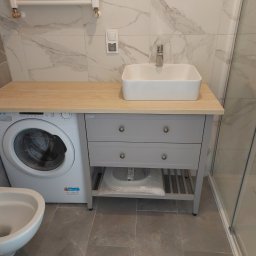 Huber Design - Nowoczesna łazienka z pralką obok szarej szafki z umywalką na drewnianym blacie, widoczna kabina prysznicowa i marmurowe płytki na ścianach.