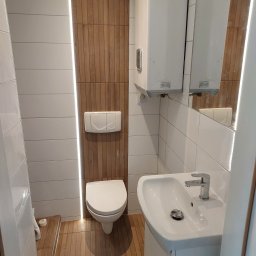 Huber Design - Nowoczesna łazienka z białymi i drewnopodobnymi płytkami, podświetleniem LED, wiszącą toaletą i umywalką na szafce, uzupełniona bojlerem Ariston i lustrem.