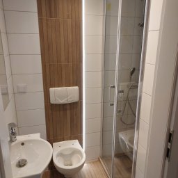 Huber Design - Nowoczesna łazienka po remoncie, z kabiną prysznicową ze szklanymi drzwiami, umywalką i podwieszaną toaletą, wykończona białymi płytkami i panelami imitującymi drewno, oświetlona ledowym paskiem.