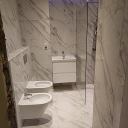 Huber Design - Wykończona łazienka z marmurowymi płytkami, wiszącą miską WC i bidetem, białą szafką umywalkową oraz kabiną prysznicową ze szklaną ścianką; widoczny fragment ściany przed wykończeniem.