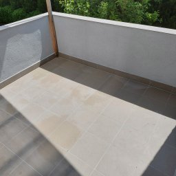 Huber Design - Wykończony balkon z jasnymi, kwadratowymi płytkami podłogowymi i jasnoszarymi ścianami z fakturą, widok z góry z cieniem padającym na podłogę.