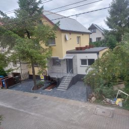 Huber Design - Żółty dom jednorodzinny z szarym dachem, nową elewacją z elementami dekoracyjnymi i podjazdem wyłożonym kostką brukową w dwóch odcieniach szarości, widok z góry.