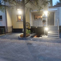 Huber Design - Oświetlony dom jednorodzinny z kostki brukowej, schodami z metalową barierką i dekoracyjnym kołem od wozu na elewacji, widoczny mężczyzna na ganku.