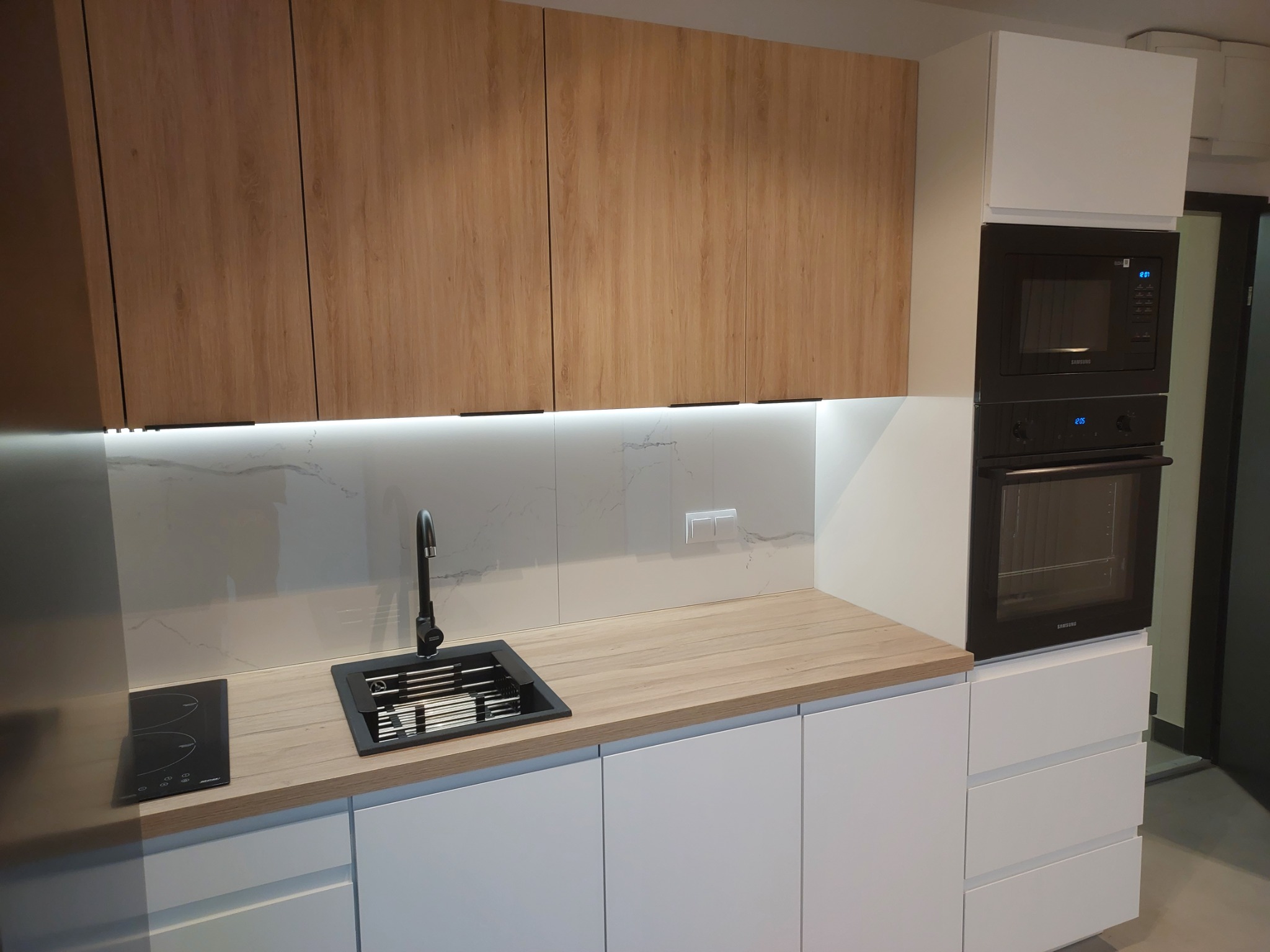 Nowoczesna kuchnia z drewnianymi szafkami górnymi, białymi szafkami dolnymi, czarnym zlewem i płytą indukcyjną, oświetleniem LED pod szafkami i zabudowanym piekarnikiem z mikrofalówką.