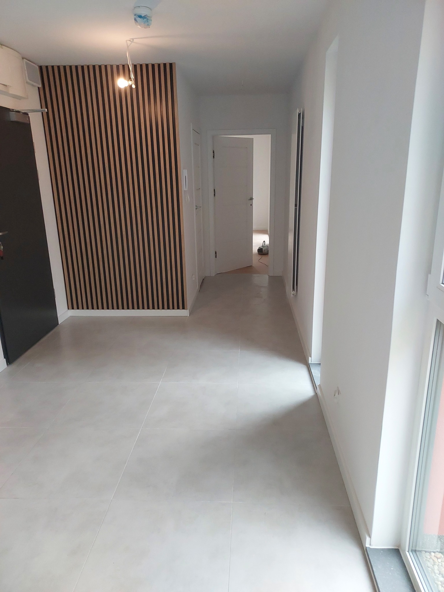 Jasne wnętrze mieszkania po remoncie, z szarą podłogą z płytek, drewnianą ścianą z paneli pionowych, widok na otwarte drzwi do kolejnego pomieszczenia i duże okno z prawej strony.