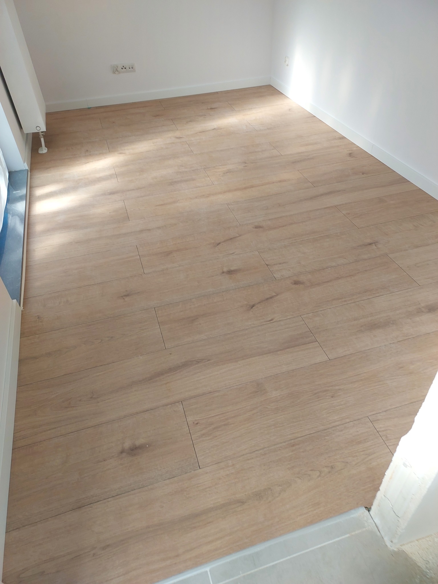 Jasny parkiet ułożony w pustym pokoju z białymi ścianami i listwami przypodłogowymi, widoczne gniazdka elektryczne i fragment grzejnika.