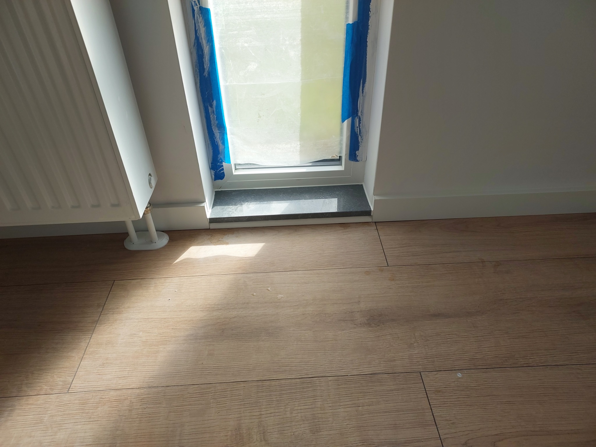 Fragment wnętrza z ułożonymi panelami podłogowymi, grzejnikiem i oknem zabezpieczonym taśmą malarską.