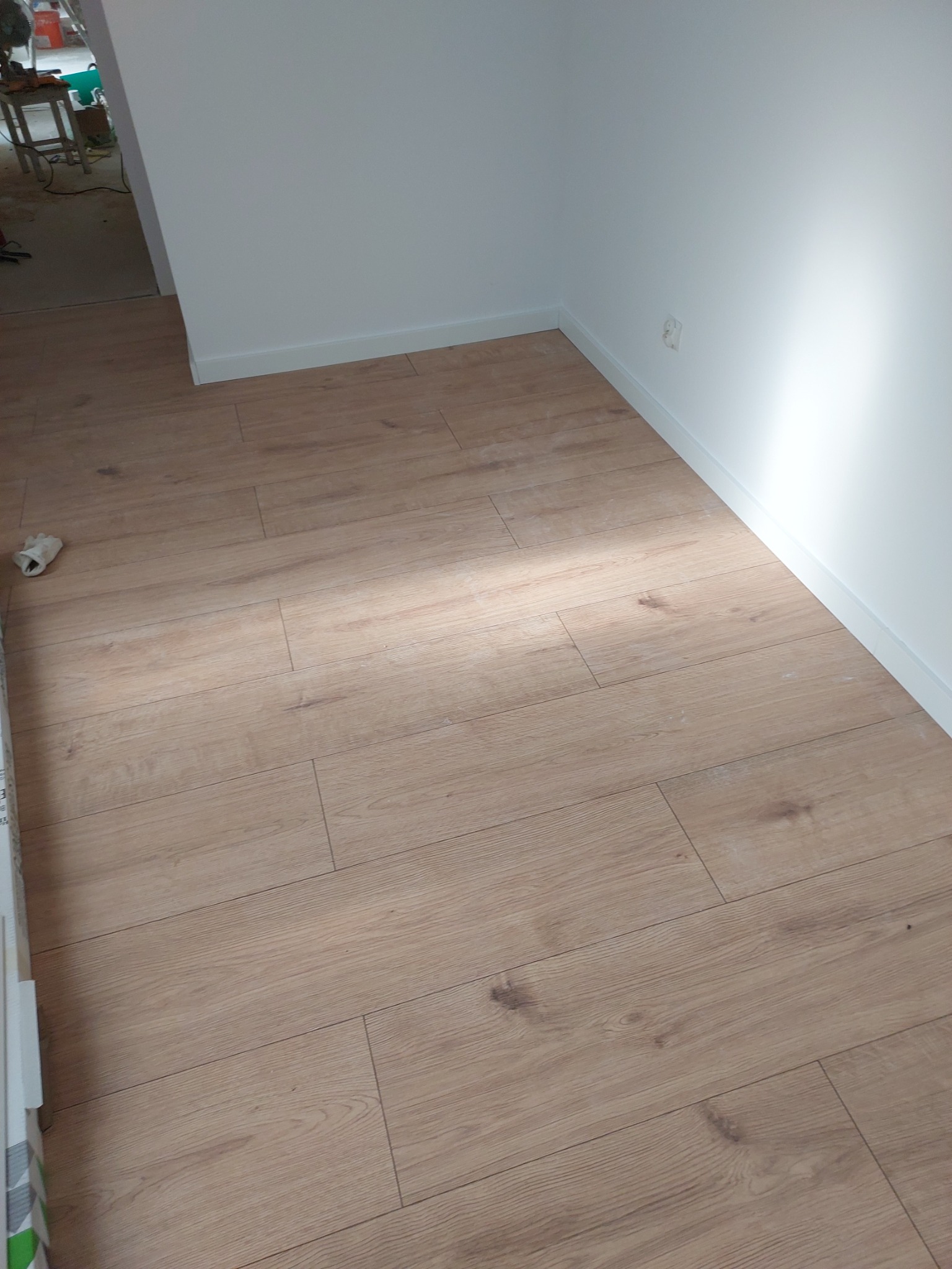 Jasna, drewnopodobna podłoga ułożona w pokoju z białymi ścianami i listwami przypodłogowymi, widoczny fragment kolejnego pomieszczenia z narzędziami i materiałami budowlanymi.