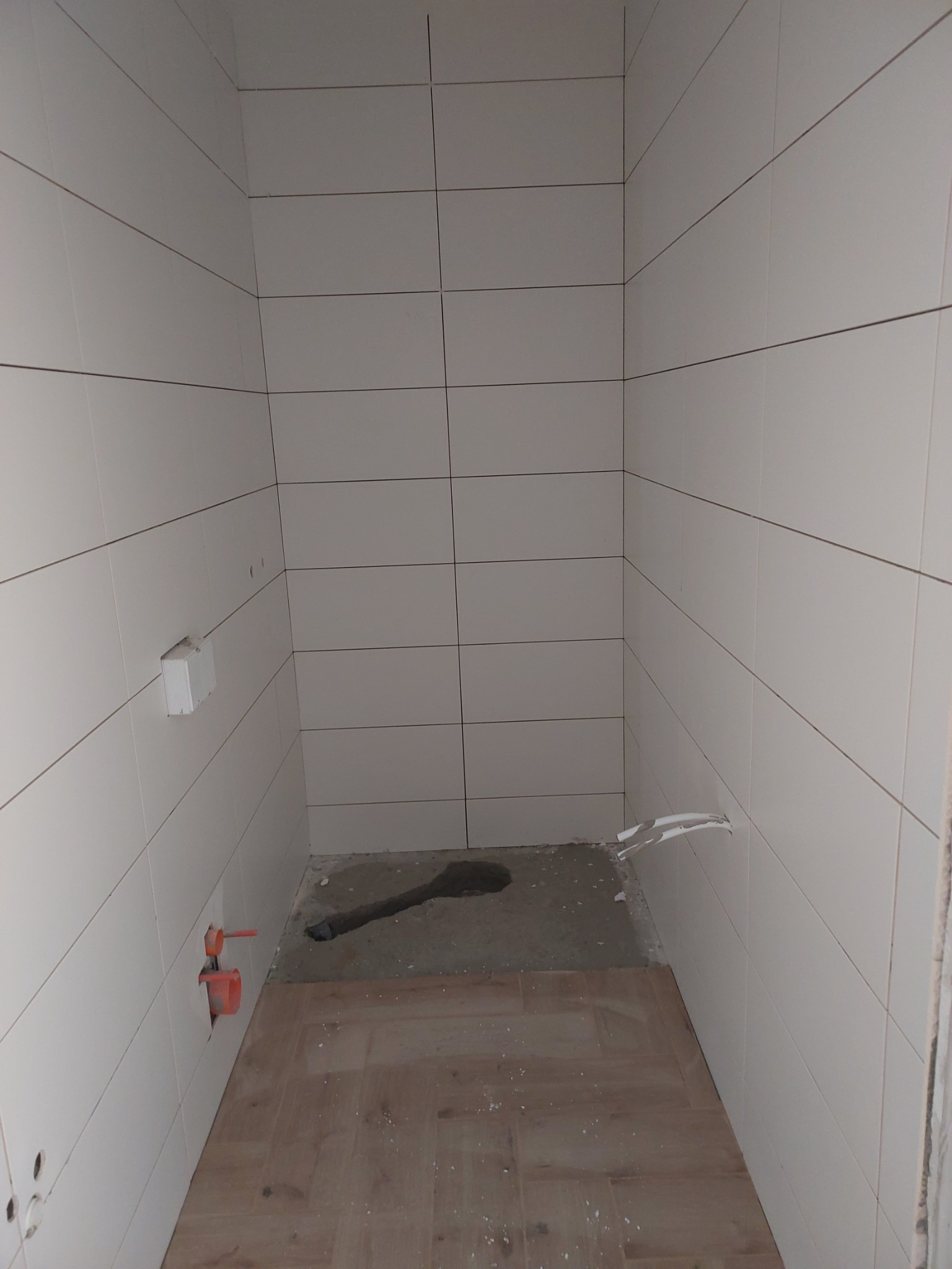 Wnętrze łazienki w trakcie prac wykończeniowych; widoczne białe płytki na ścianach, częściowo ułożona podłoga z paneli, wystające rury instalacji wodnej i kanalizacyjnej, oraz pozostałości zaprawy...