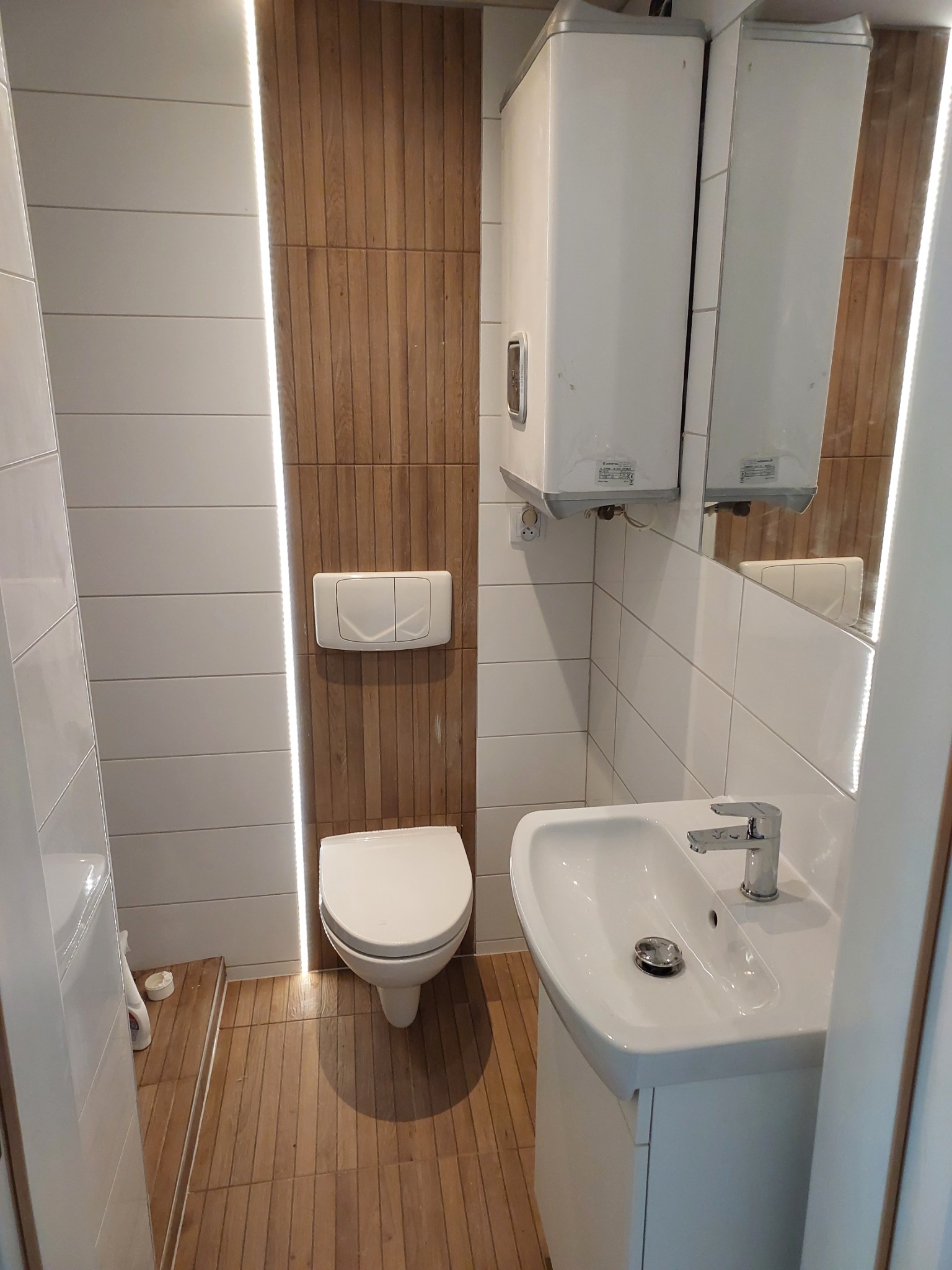 Nowoczesna łazienka z białymi i drewnopodobnymi płytkami, podświetleniem LED, wiszącą toaletą i umywalką na szafce, uzupełniona bojlerem Ariston i lustrem.