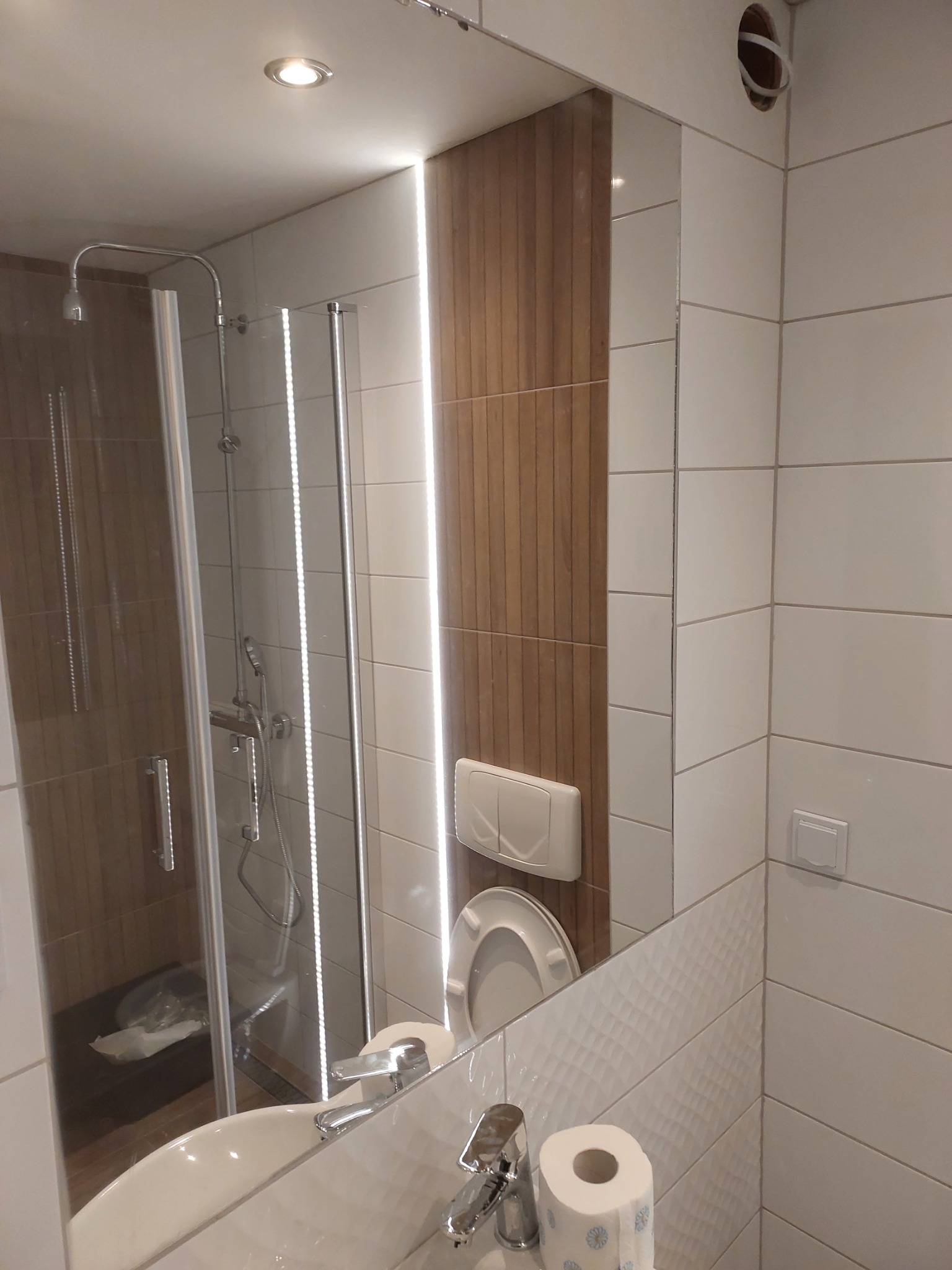 Wykończona łazienka z kabiną prysznicową, umywalką i toaletą, widoczna w lustrze, z elementami drewnopodobnymi i oświetleniem LED, z niedokończonym otworem na wentylację.