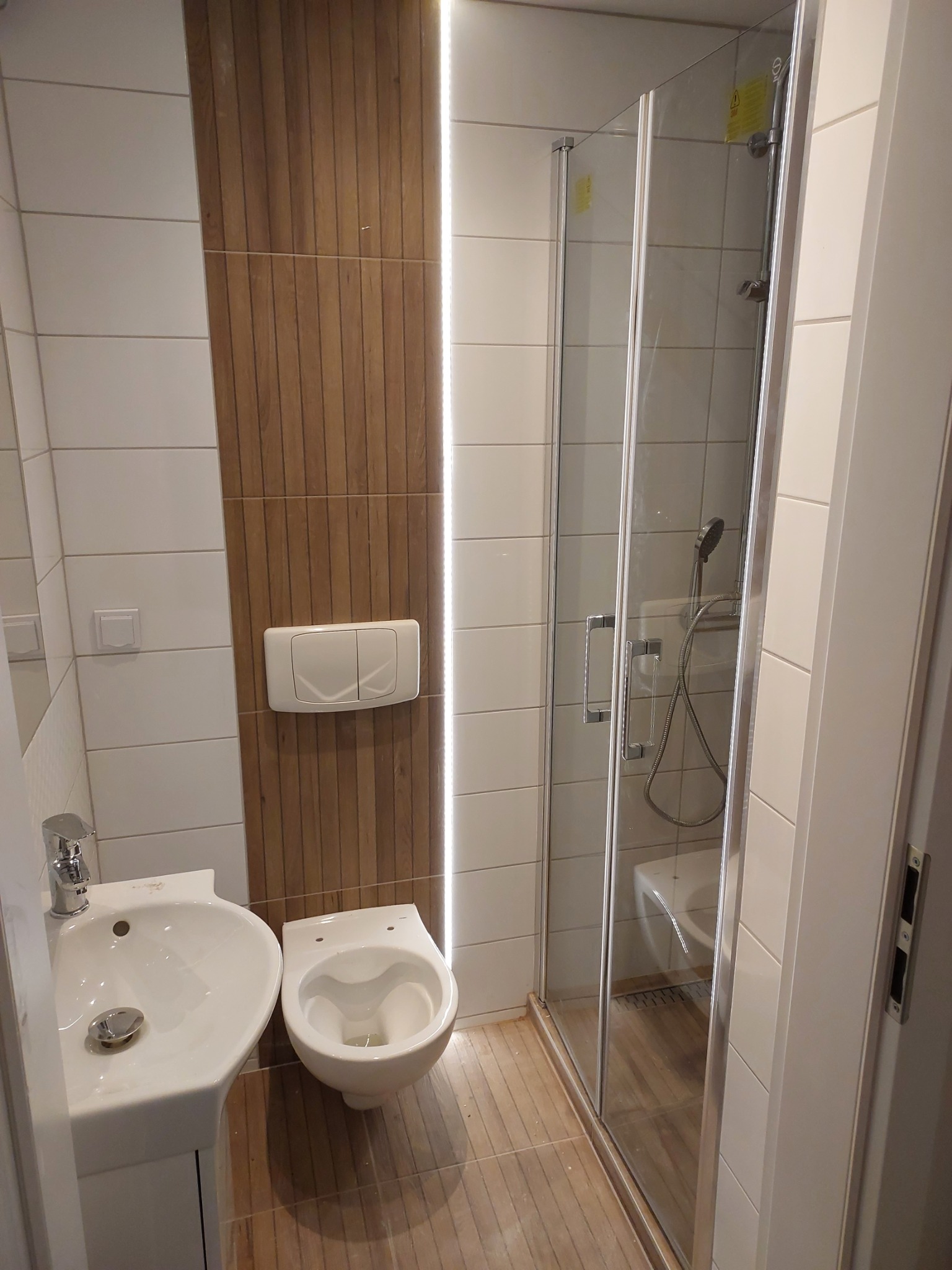 Nowoczesna łazienka po remoncie, z kabiną prysznicową ze szklanymi drzwiami, umywalką i podwieszaną toaletą, wykończona białymi płytkami i panelami imitującymi drewno, oświetlona ledowym paskiem.