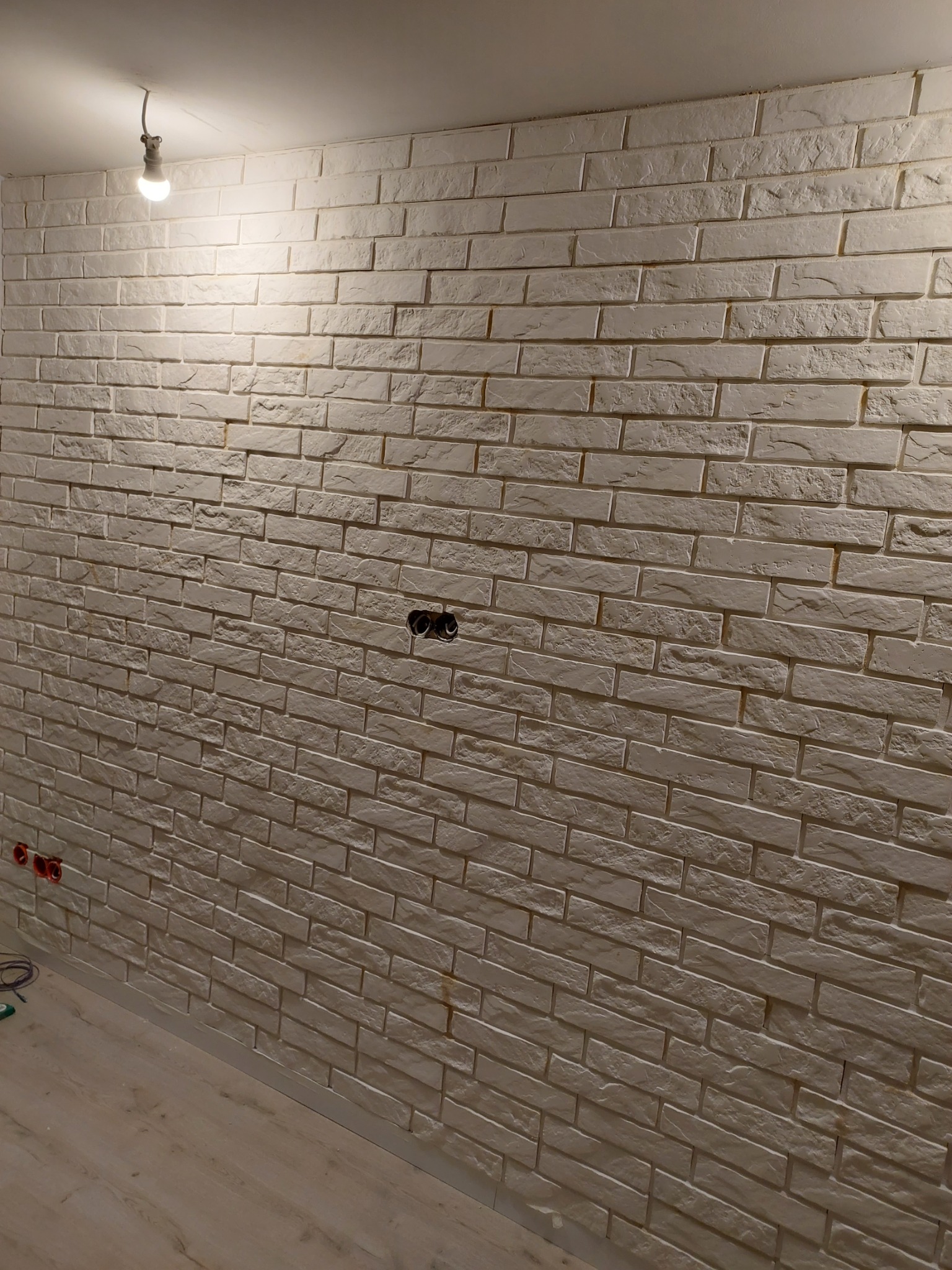 Ściana wyłożona dekoracyjną cegłą w odcieniach bieli, widoczne gniazdka elektryczne bez osprzętu, podłoga z paneli, surowa żarówka na kablu.
