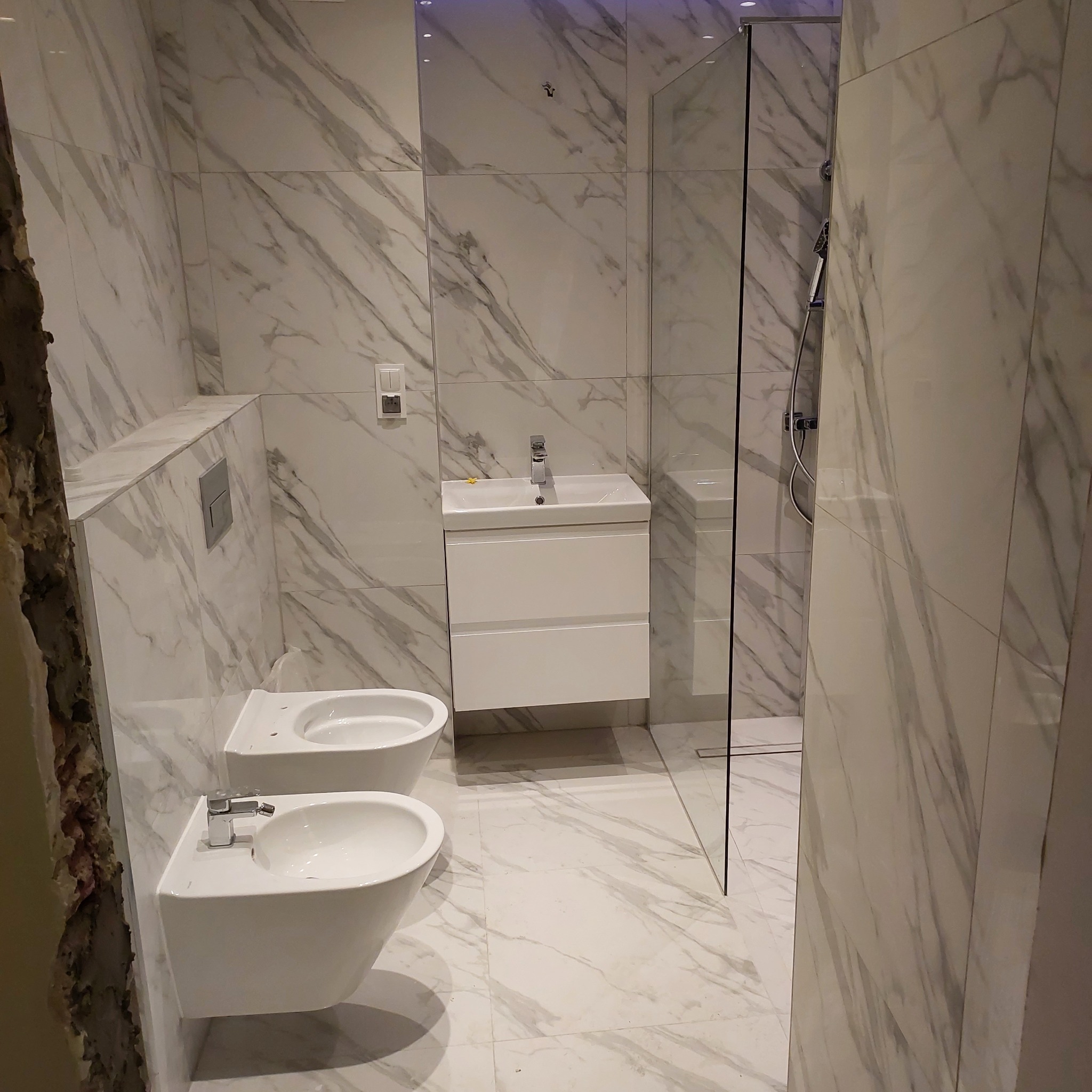 Wykończona łazienka z marmurowymi płytkami, wiszącą miską WC i bidetem, białą szafką umywalkową oraz kabiną prysznicową ze szklaną ścianką; widoczny fragment ściany przed wykończeniem.
