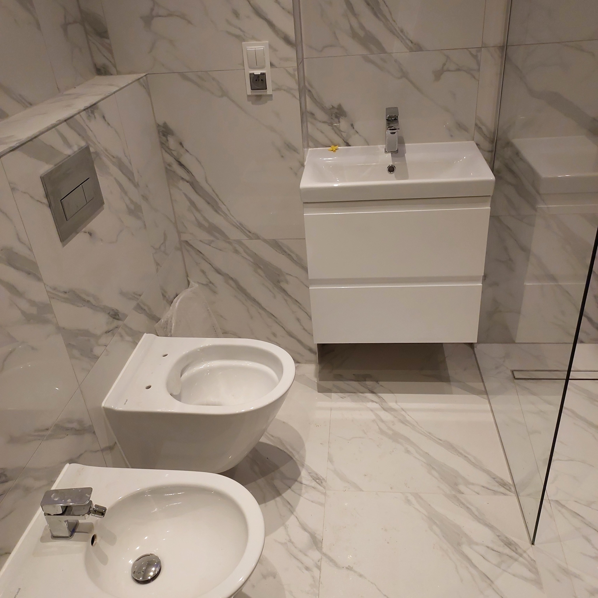 Nowoczesna łazienka z marmurowymi płytkami, wiszącą toaletą, bidetem i umywalką z szafką w kolorze białym.