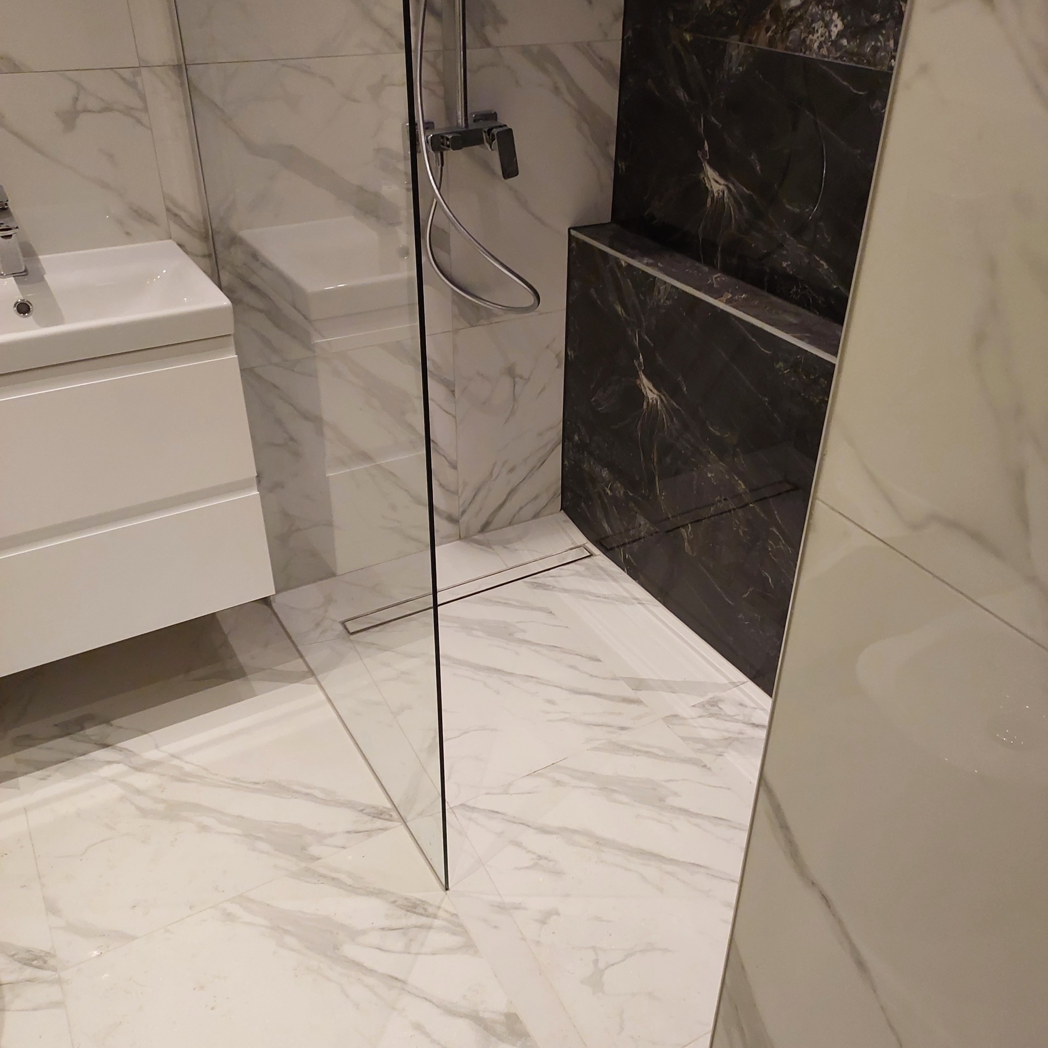 Nowoczesna łazienka z kabiną prysznicową typu walk-in, białą umywalką z szafką i marmurowymi płytkami w odcieniach bieli i czerni, widoczny odpływ liniowy.