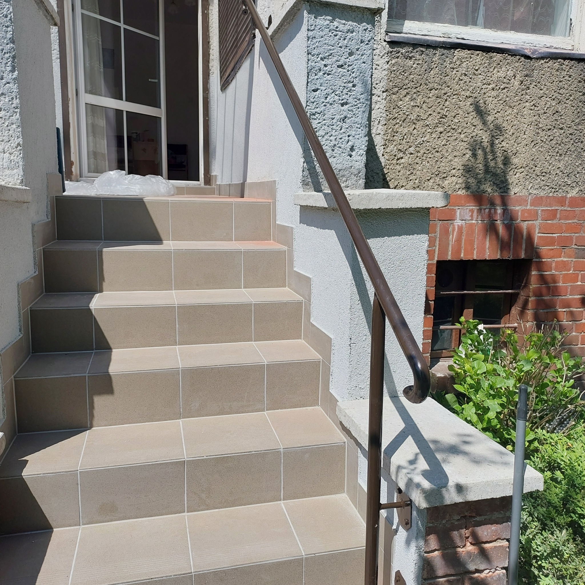 Wykończone schody wejściowe do domu, wyłożone beżowymi płytkami ceramicznymi, z brązową metalową poręczą, widoczne detale elewacji i roślinność