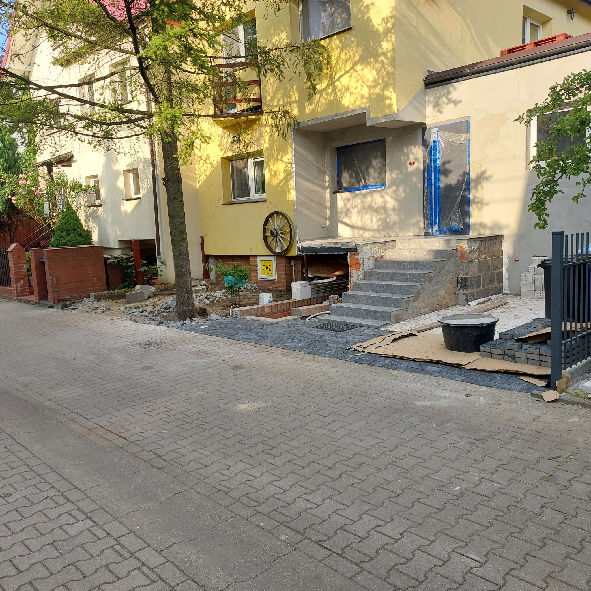 Remont wejścia do budynku: nowe schody z szarej kostki, przygotowane bloczki betonowe, wiadro z zaprawą, folia ochronna na drzwiach wejściowych, kostka brukowa na chodniku, widoczne elementy...