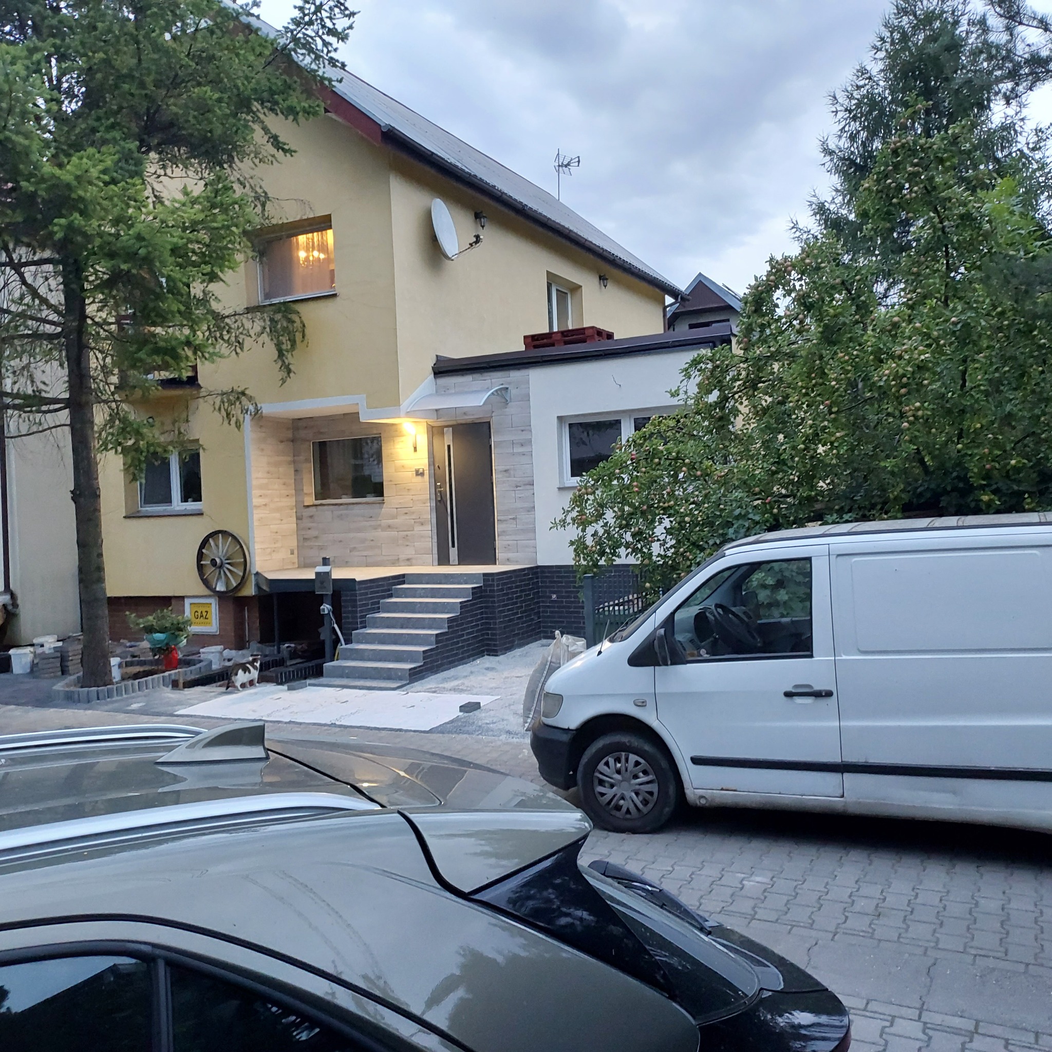 Żółty dom jednorodzinny po renowacji elewacji i schodów wejściowych, z zaparkowanym białym vanem i szarym samochodem osobowym na pierwszym planie, widoczny kot przy wejściu.